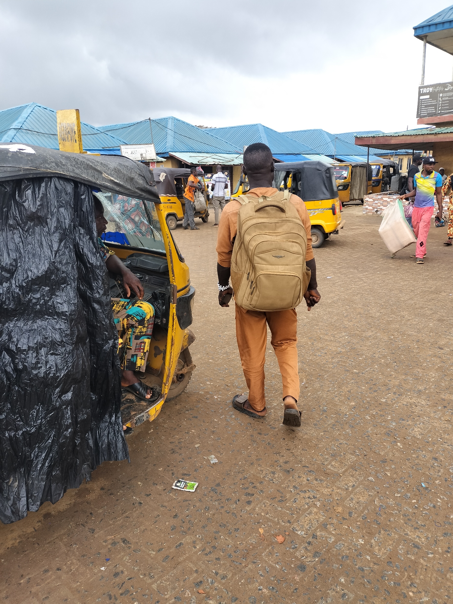 In a bustling street scene in Ikorodu, Nigeria, a man with a large khaki backpack is walking away from the camera. He is wearing matching brown pants and a long-sleeved shirt with brown sandals. To his left, a yellow auto rickshaw, partially obscured by a black plastic sheet, has a driver wearing a colorful shirt and patterned pants. Scattered trash is on the ground, and in the background, several more yellow auto rickshaws are present. People are walking around in the background, and buildings with blue roofs line the street. A sign is visible in the background with the word "TROY" on it.