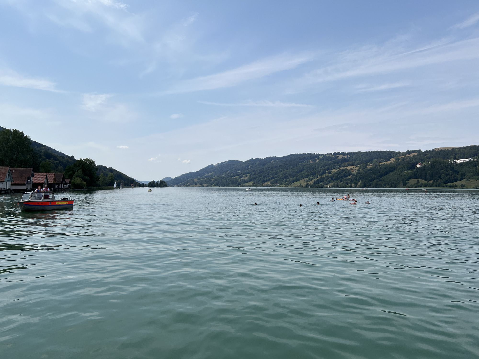 This image depicts a scenic view of the Alpsee, a lake located in Immenstadt im Allgäu, Germany. The Alpsee is renowned for its crystal-clear water and the picturesque mountain backdrop. In the foreground, the calm, turquoise waters of the lake reflect the soft, hazy sunlight. Several people are visible enjoying recreational activities on the lake, with some swimming and others relaxing on inflatable rafts. A small red and orange boat with two people on board is positioned near the left edge of the frame, close to a cluster of quaint, reddish-roofed houses that line the shore. In the distance, the lush, green slopes of the surrounding mountains rise, their peaks partially obscured by a light, wispy cloud cover. The sky above is a pale blue, with streaks of white clouds adding texture to the expansive sky. Sailboats can be seen dotted across the lake, hinting at a pleasant day for outdoor pursuits.
