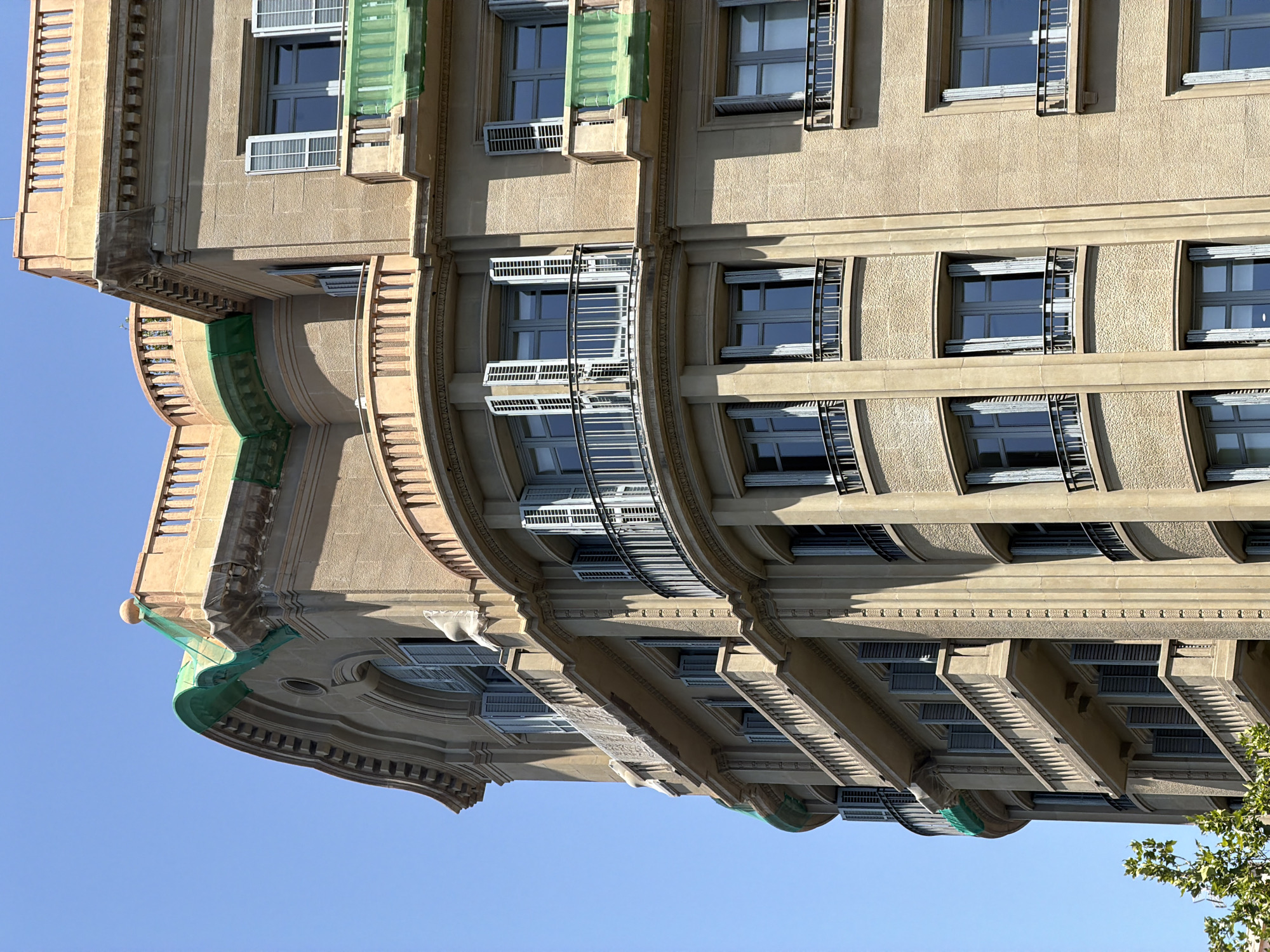 This is a low-angle, full shot of a building's facade against a clear blue sky. The building is an architectural structure with several levels, featuring repetitive window designs and decorative elements. The windows appear to be framed with white shutters and are set within light-colored walls. Some windows are open, revealing the dark interiors. The building has curved balconies with railings and ornate moldings, suggesting a classical or art deco style. A green mesh netting is visible in some sections, indicating possible construction or maintenance work. The perspective is unusual, as the image appears to be rotated, with the top of the building at the bottom of the frame. The lighting suggests it is daytime, with sunlight casting shadows on the building's surfaces, highlighting the architectural details and textures. No people or text are visible in the image. The location is identified as Barcelona, Spain.