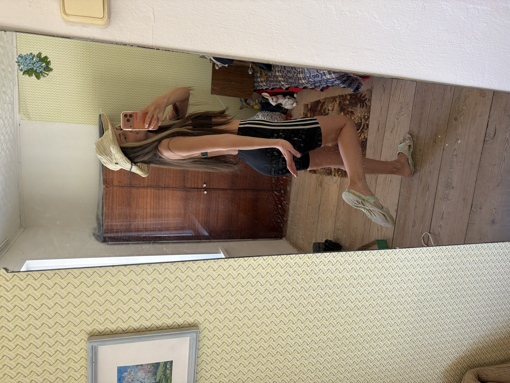 A mirror reflection shows a young woman in a straw hat, black tank top, and Adidas shorts, holding a mobile phone in front of her face. She is standing in a room with patterned wallpaper, a wooden floor, and a rug with clothing and objects strewn on it. A dresser is visible behind her in the reflection. The location in Kamyshin, Russia, is not discernible from the image.