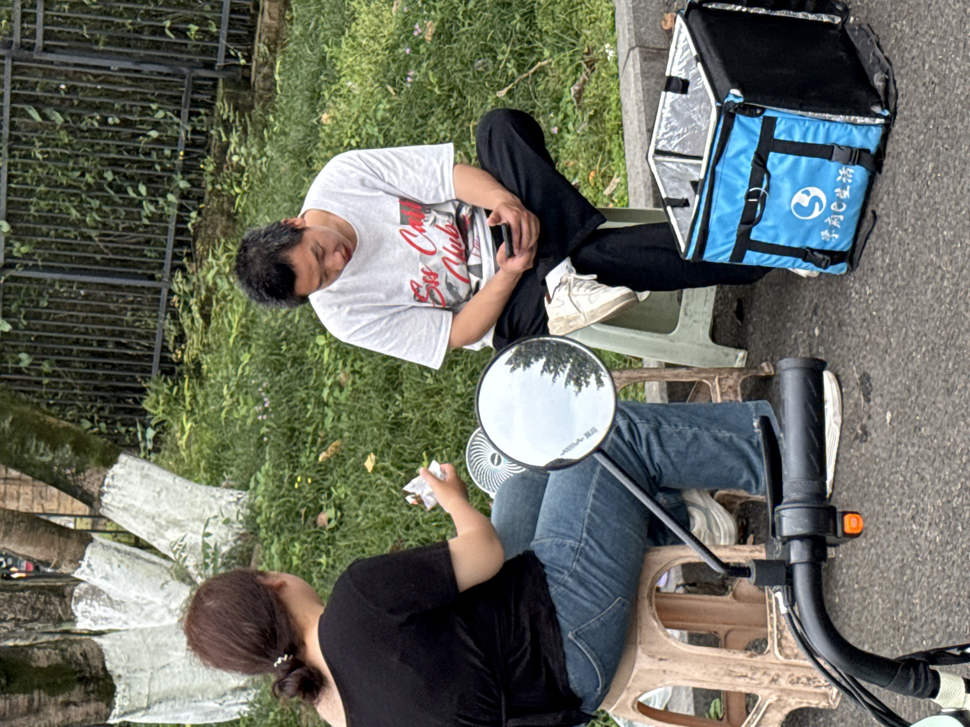 Two people are seated on small plastic stools on a street in Jiangjin, China. One person, a male, is wearing a white t-shirt and dark pants, and is looking down at a phone in his hands. Next to him is a blue delivery bag. The other person, a female, is wearing a black shirt and blue jeans. She is holding something in her hand. In front of her is the handlebar of a motorcycle with a mirror. The background includes a fence, grass, and trees.