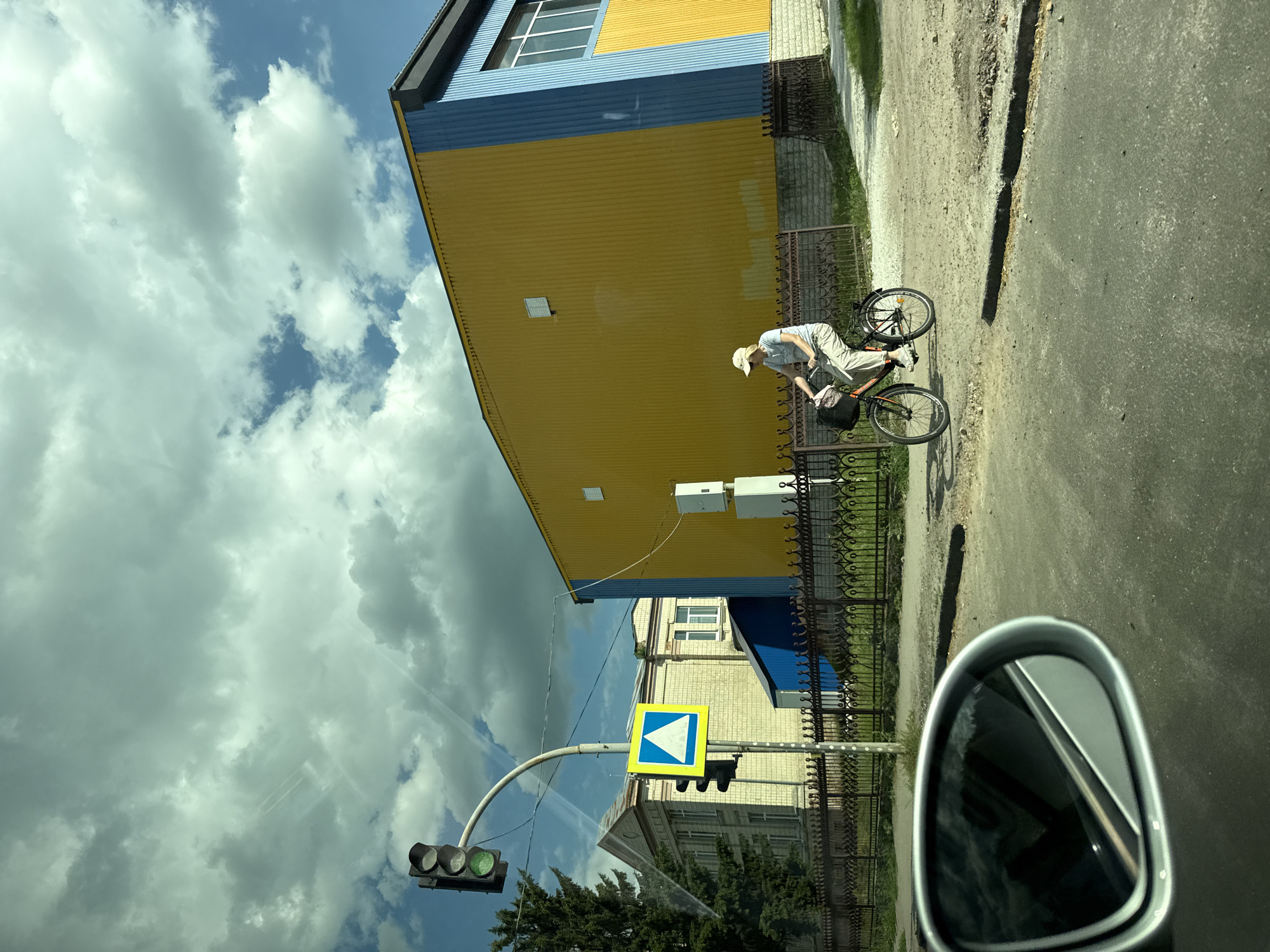 A person is riding a bicycle down a paved road, which appears to be a street in Skvyra, Ukraine. The person is wearing a light-colored hat, shirt, and pants, and is leaning forward over the handlebars. The bicycle has a black bag attached to the rear. The setting is outdoors, with a bright, cloudy sky overhead. To the left of the road, there is a building with a yellow and blue facade. A black metal fence runs along the side of the building. Further down the street, a traffic light is visible, with the green light illuminated. There is also a yellow and blue diamond-shaped road sign with a white arrow pointing to the right. The reflection in the car's side mirror shows the road and sky. The image appears to have been taken during the daytime, likely in the late morning or early afternoon, given the brightness of the sunlight filtering through the clouds. The overall impression is a casual scene of a person cycling on a day with mixed weather. There is no visible text in the image, other than what might be on the road sign, which is a directional arrow.