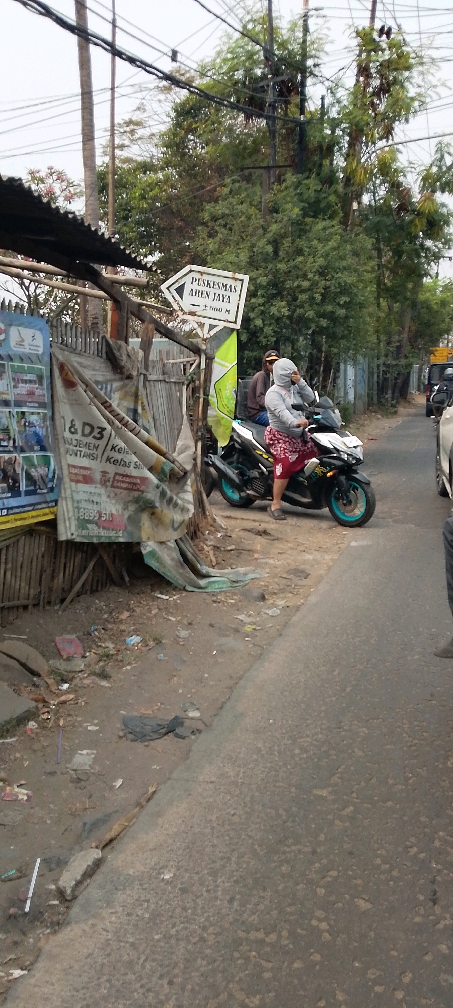 In Bekasi, Indonesia, an outdoor scene unfolds on the side of a road. A white directional sign pointing to the right reads "PUSKESMAS AREN JAYA ± 800 M". To the left of the sign, a makeshift structure of wood and corrugated metal stands, partially covered by a tattered white and green banner. The banner displays text, including what appears to be "D3 MANAGEMENT" and a phone number "1-8899 511". A person wearing a grey hoodie and patterned red skirt rides a black motorcycle with teal accents. Behind them, another individual is seated on the same motorcycle, wearing a brown jacket and a cap. The ground is dirt and asphalt, littered with debris, including plastic bags and what looks like discarded bamboo. In the background, lush green trees and utility poles with a complex network of wires dominate the landscape. A glimpse of a white car and another motorcycle further down the road suggests ongoing traffic. The overall atmosphere is that of a busy, somewhat unkempt urban environment.