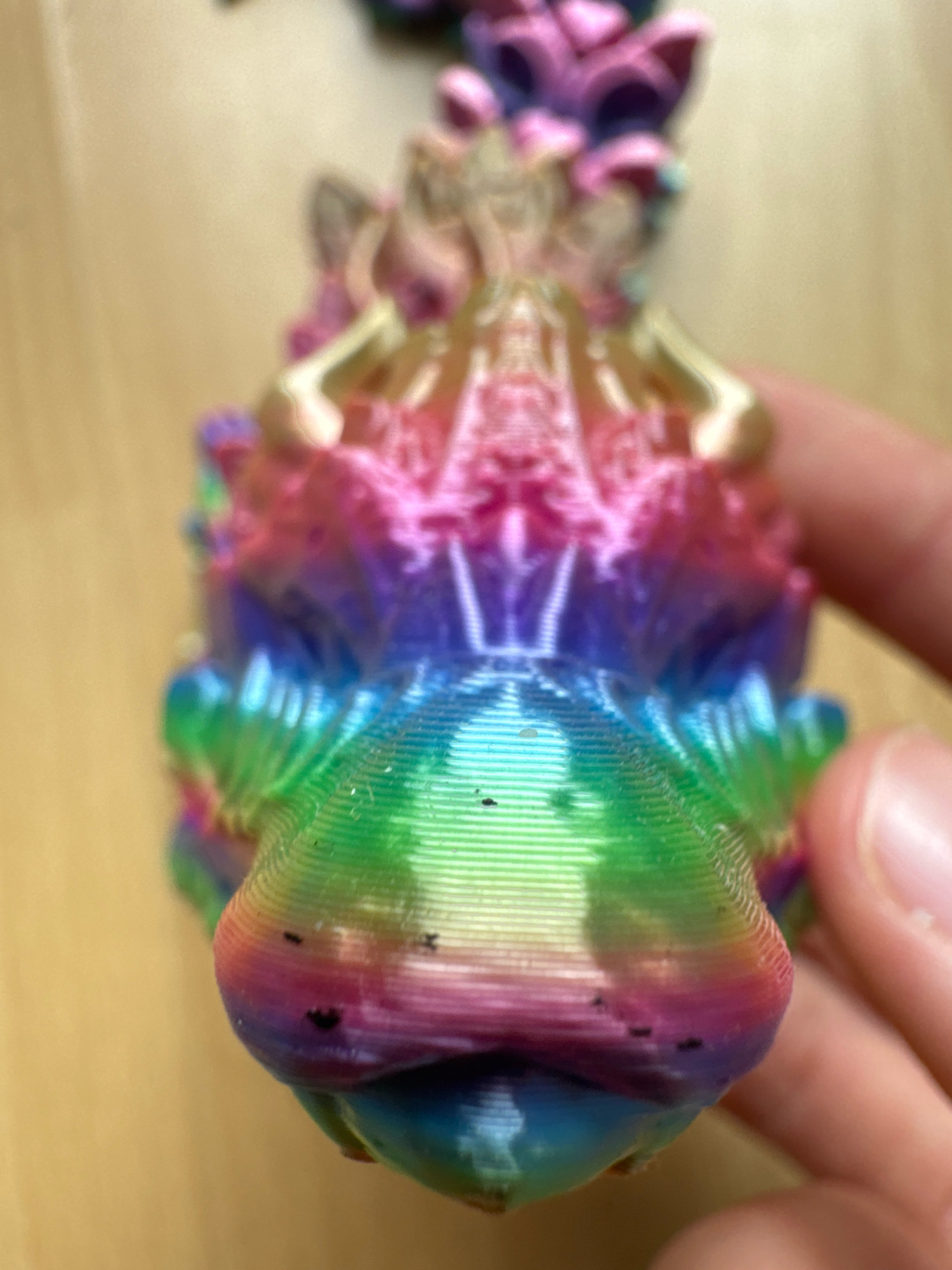 A close-up, angled shot focuses on a rainbow-colored, 3D-printed dragon figurine held by a human hand. The figurine displays distinct layer lines characteristic of 3D printing. Its head, predominantly green and purple, shows small black specks, possibly dust or imperfections. The background consists of a blurry, light brown surface, likely a wooden table. The image lacks any discernible urban elements or connection to the city of Überherrn, Germany.