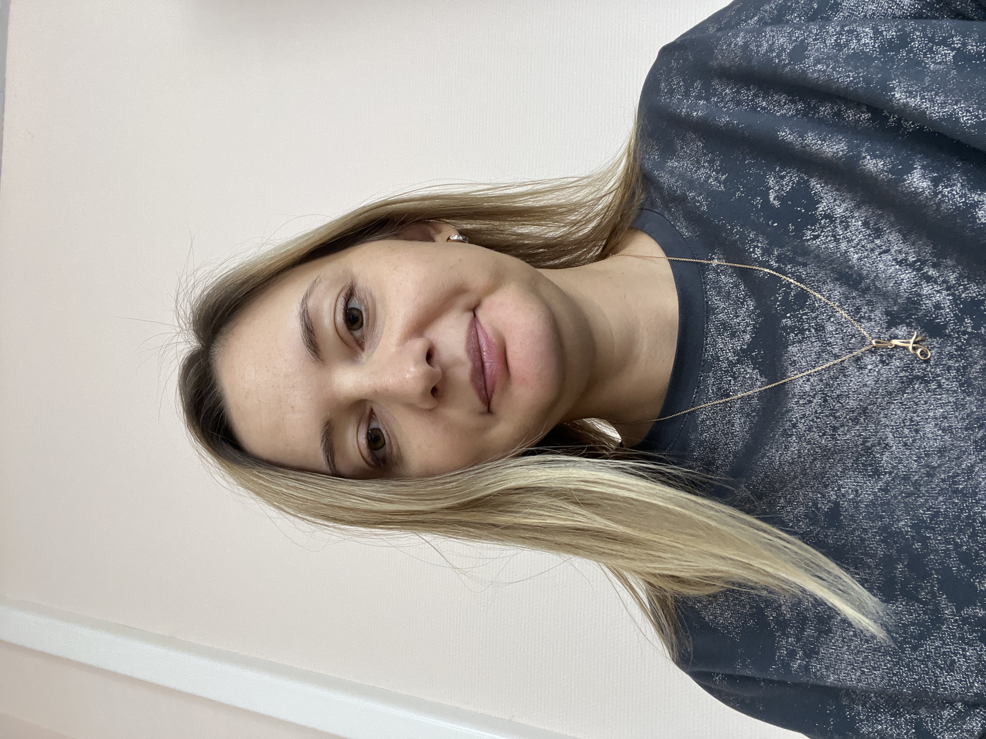 A medium shot, front-facing selfie shows a woman with shoulder-length blonde hair and brown eyes. She is smiling gently and looking directly at the camera. She is wearing a dark gray crewneck sweatshirt with a textured, shimmering pattern. A thin gold chain with a charm hangs around her neck. She has a small hoop earring in her left ear. The background appears to be a plain, light-colored wall with a white vertical trim on the left side of the frame. There is no indication of the specific location in Volgograd, Russia.