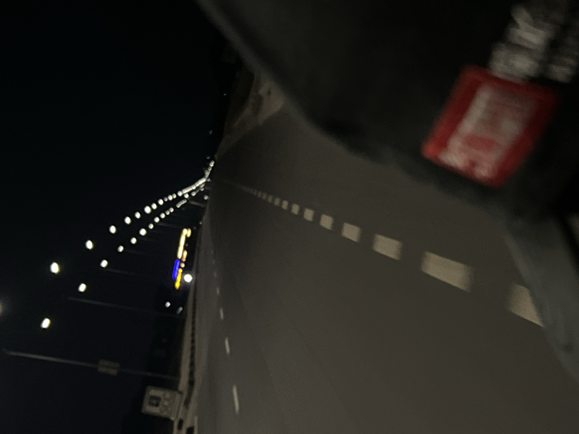 The image depicts a dark, blurry view of a bridge at night. The bridge has lights lining the side, and there are dashed white lines indicating lanes of traffic. A portion of an object with a red label is visible in the foreground, suggesting it is near the camera.
I cannot determine if the bridge is located in the city of Dam Camp, Nigeria, due to the lack of location cues.