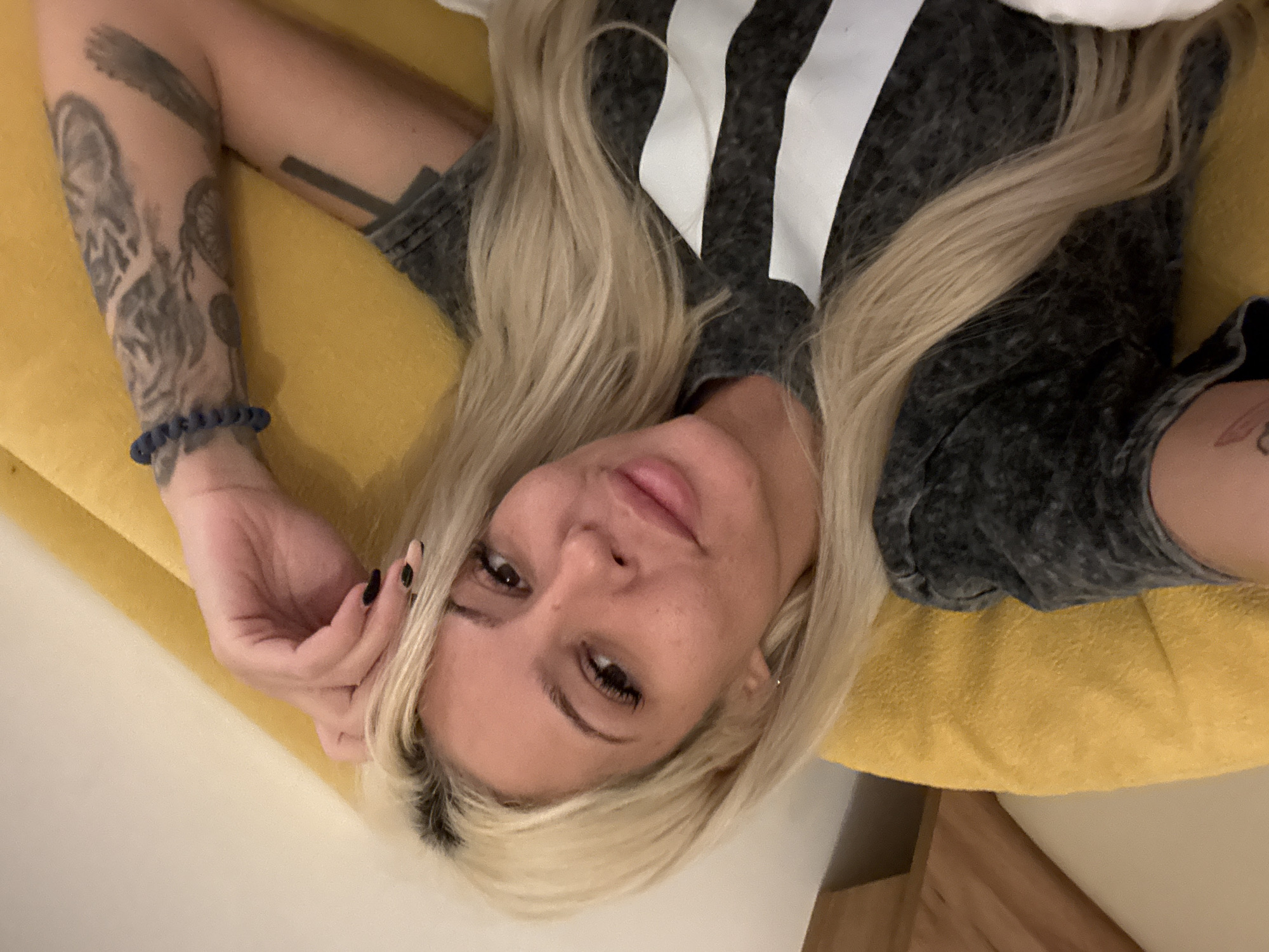 The image, taken from an overhead perspective, depicts a woman with long blonde hair and darker roots, lying on a vibrant yellow surface. Her head is slightly tilted, and she is gazing directly at the camera with a gentle, relaxed smile. She has full lips, dark eyes, and prominent eyelashes.
She is wearing a dark grey t-shirt featuring bold white vertical stripes across the upper part. Her left arm is visible, adorned with several tattoos, including an intricate design on her forearm that appears to be an animal head, possibly a tiger or lion. A blue beaded bracelet is worn on her left wrist, and her fingernails are painted black.
The woman is positioned on a soft, textured bright yellow material, likely a pillow, cushion, or upholstered furniture. The background is a plain, light-colored surface, suggesting an indoor setting. A small portion of light wooden furnishing is visible in the bottom right corner. The lighting is soft and even, indicating a well-lit indoor environment. No visible text or brand logos are discernible. The scene is set indoors in Sofia, Bulgaria.