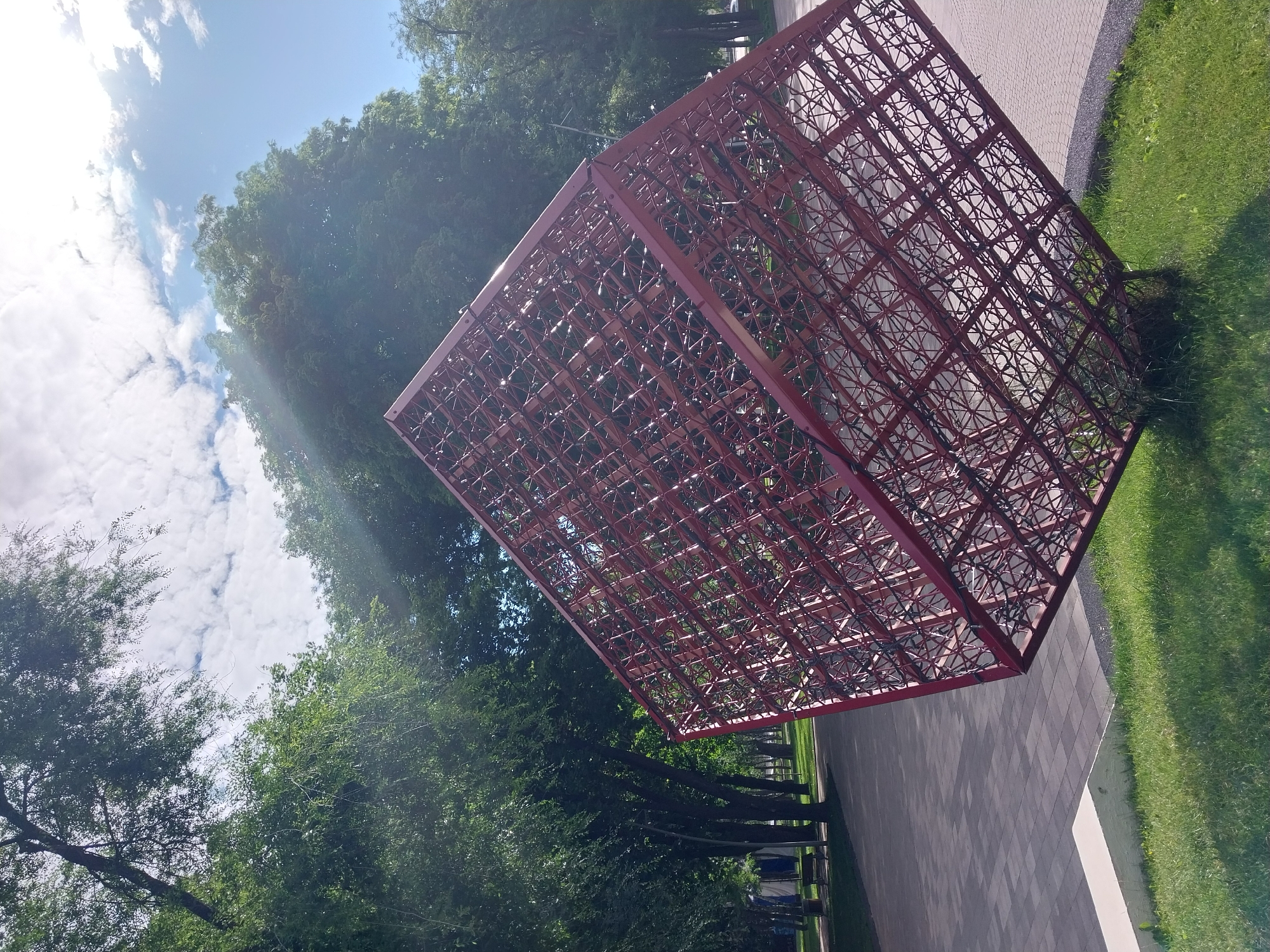 The image depicts an abstract red metal sculpture shaped like a large cube, tilted on its side, situated in a park area with a paved walkway and green grass. The sculpture is intricately constructed with a grid-like pattern of intersecting metal bars. In the background, lush green trees form a dense canopy, and the sky is bright blue with scattered white clouds, indicating daytime. Sunlight streams through the trees, creating lens flare effects. The location is identified as Dnipro, Ukraine. No people are visible in the scene.