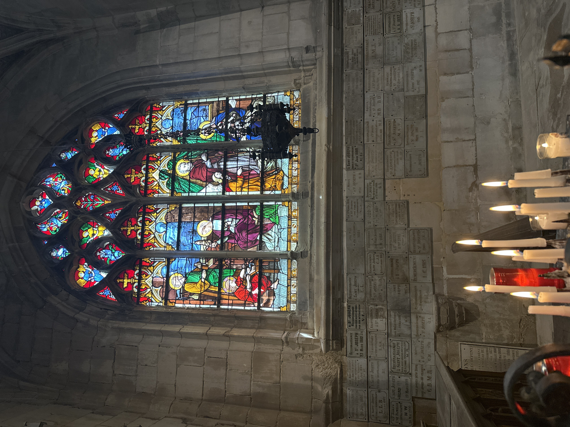 This image depicts a view inside a church in Dieppe, France, featuring a large, ornate stained-glass window and a wall covered in commemorative plaques. The stained-glass window is divided into multiple panels, each depicting figures in richly colored robes, likely religious scenes or saints, with intricate patterns and vibrant blues, reds, yellows, and greens. A dark, wrought-iron chandelier hangs in front of the window. Below the window, the wall is densely covered with numerous rectangular, light-colored plaques, each bearing inscribed text, presumably memorials to deceased individuals. In the foreground, several lit candles of varying sizes and colors, including a large red candle in a glass holder, are arranged on a surface, casting a warm glow. The overall environment suggests a solemn and historical interior, consistent with a church in Dieppe, a coastal city in Normandy, France, known for its historical architecture and religious sites. The wall texture and the stonework visible on the left and above the window indicate a traditional church construction.