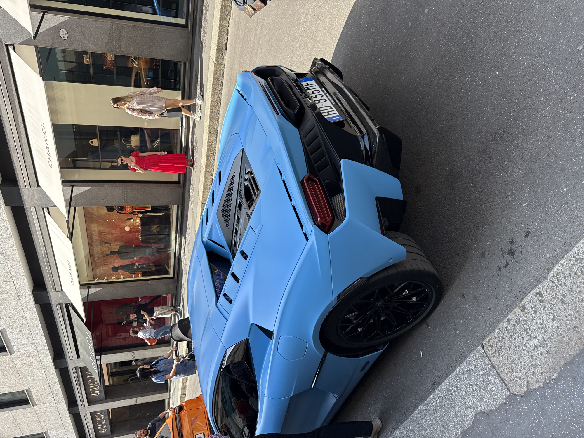 The image shows a light blue Lamborghini parked on a street next to high-end fashion stores. A Chanel store is visible on the left, featuring mannequins in the window display. Further down the street, a Gucci store is also identifiable. Several pedestrians are present, some are window shopping, others are walking along the sidewalk. The car has an Italian license plate, "HD-856ME", and the plate includes text "LAMBORGHINI MILANO." The scene is located in Milan, Italy.