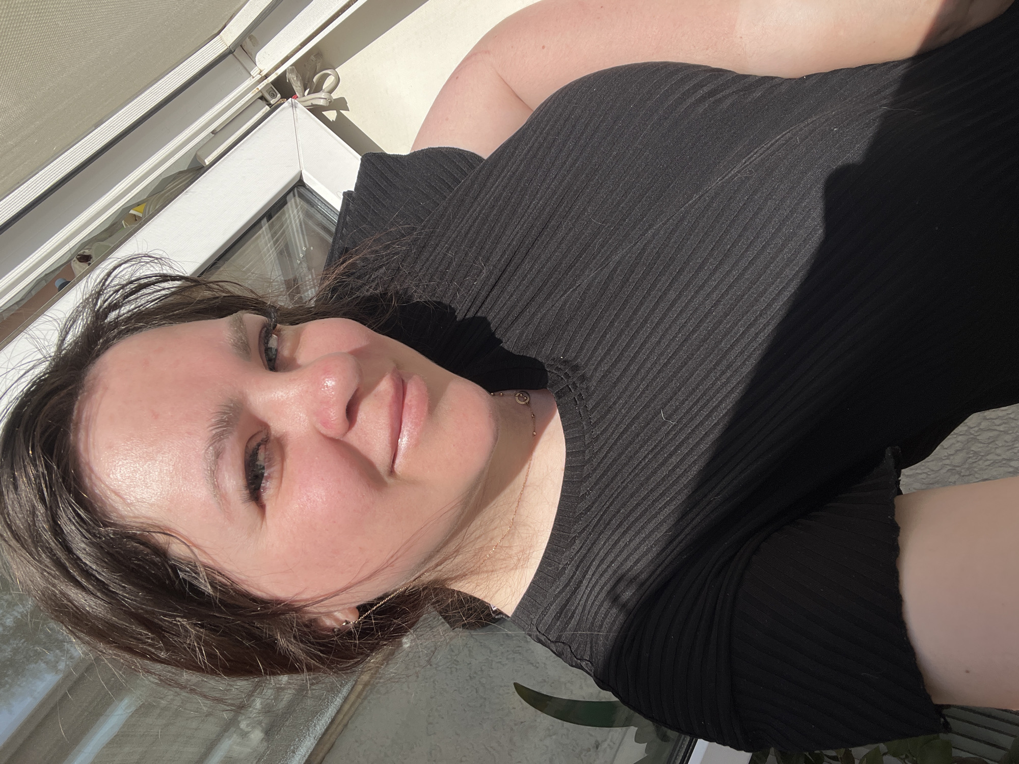 A woman with fair skin and brown hair is taking a selfie. She is wearing a black ribbed top. She is facing the camera with a slight tilt of her head. The background includes a window with a white frame and a white window shade. A plant is partially visible in the lower right corner of the frame. The location is not discernible.