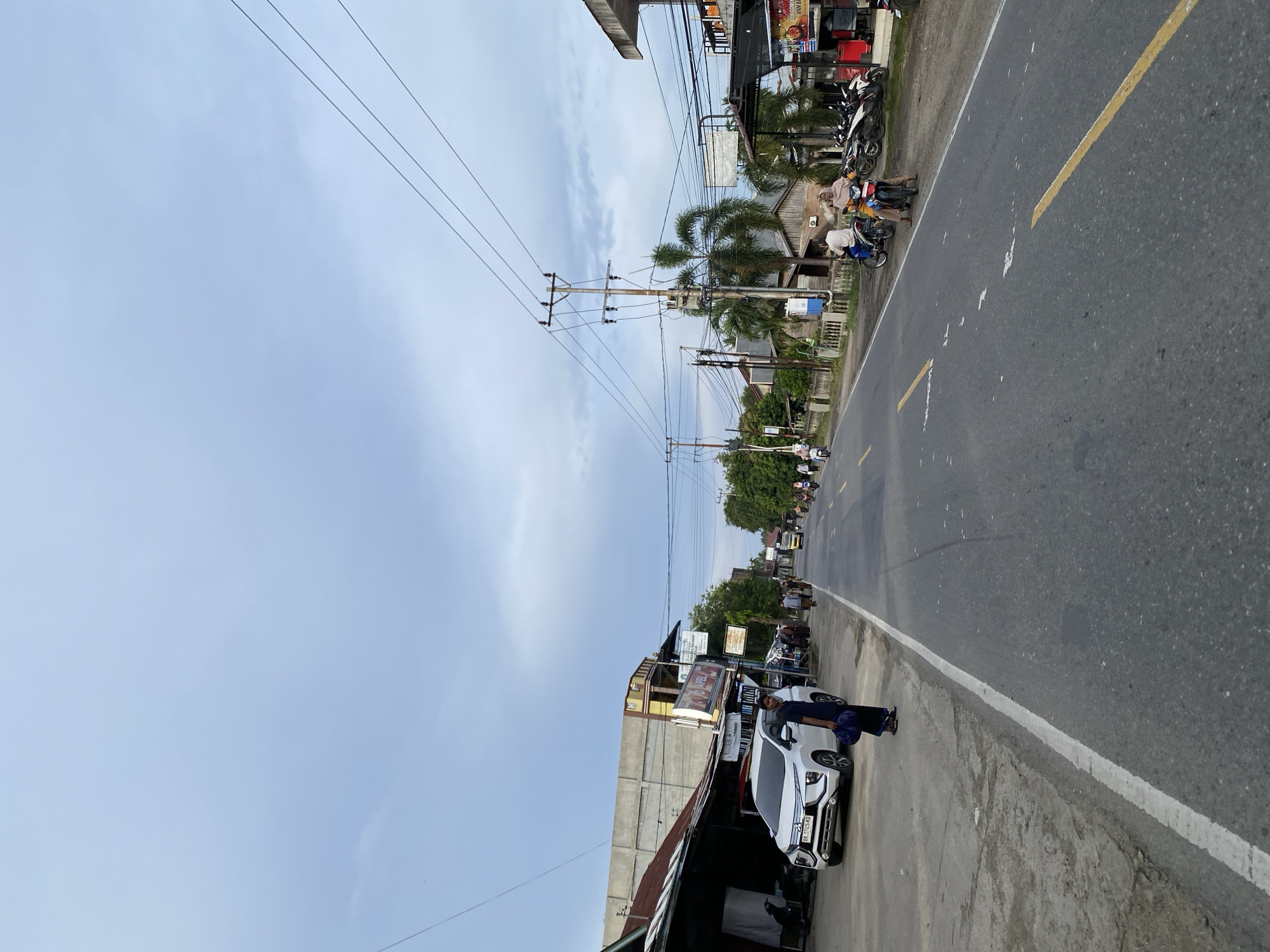 The image depicts a street scene in Cottrueng, Indonesia, under a cloudy sky. The perspective is from the side of the road, looking forward. On the left side of the road, there are buildings with corrugated metal roofs and concrete structures, one of which has a sign that reads "AKEL". A white SUV is parked on the left shoulder, and a person in dark clothing is standing near it. Further down the road, there are more buildings, palm trees, and utility poles with numerous electrical wires.
The road itself is asphalt with yellow lane markings. Several motorcycles are present. In the foreground on the right side, a person on a motorcycle is visible. Further down the road, more people on motorcycles and a yellow truck are traveling. The overall atmosphere suggests a typical day in a moderately developed area, with a mix of commercial buildings and residential structures lining the main thoroughfare.