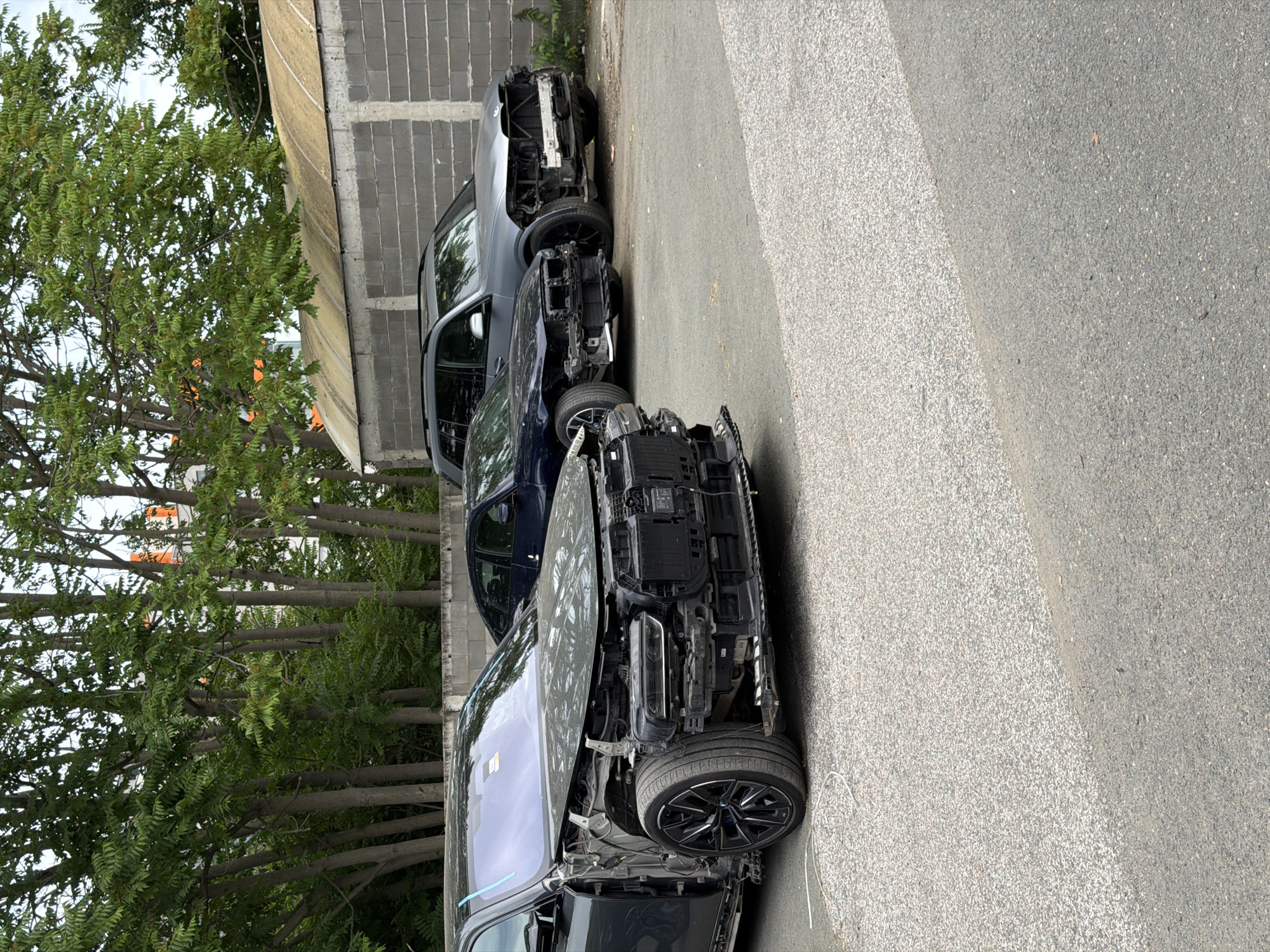 The image displays a scene with two damaged cars parked on an asphalt road. The cars are positioned next to a gray, weathered building with a concrete block facade and a slanted, yellowish roof. The building is flanked by a row of tall, slender trees with lush green foliage.
The car in the foreground is predominantly black and appears to have suffered significant damage to its front end. Its hood is open, revealing a dissembled engine and various mechanical components. The front bumper is missing, and the headlights are exposed. The wheels are large, black, and sporty, with the front wheel on the right side clearly visible.
Behind the black car is a dark blue car, also appearing to be damaged, though less severely than the first. Its front end is also partially obscured or damaged, suggesting a similar incident.
The setting appears to be outdoors, with the bright lighting suggesting daytime. The sky is not visible in the frame. The ground is paved with asphalt, and there's a distinct line separating a lighter-colored, possibly gravelly section of the road from the darker asphalt. A thin white wire or cable lies on the ground near the front of the black car.
There is no visible text in the image. The overall impression is one of damaged vehicles in an urban or semi-urban setting, possibly after an accident or awaiting repair or dismantling. The scene conveys a sense of disrepair and neglect.