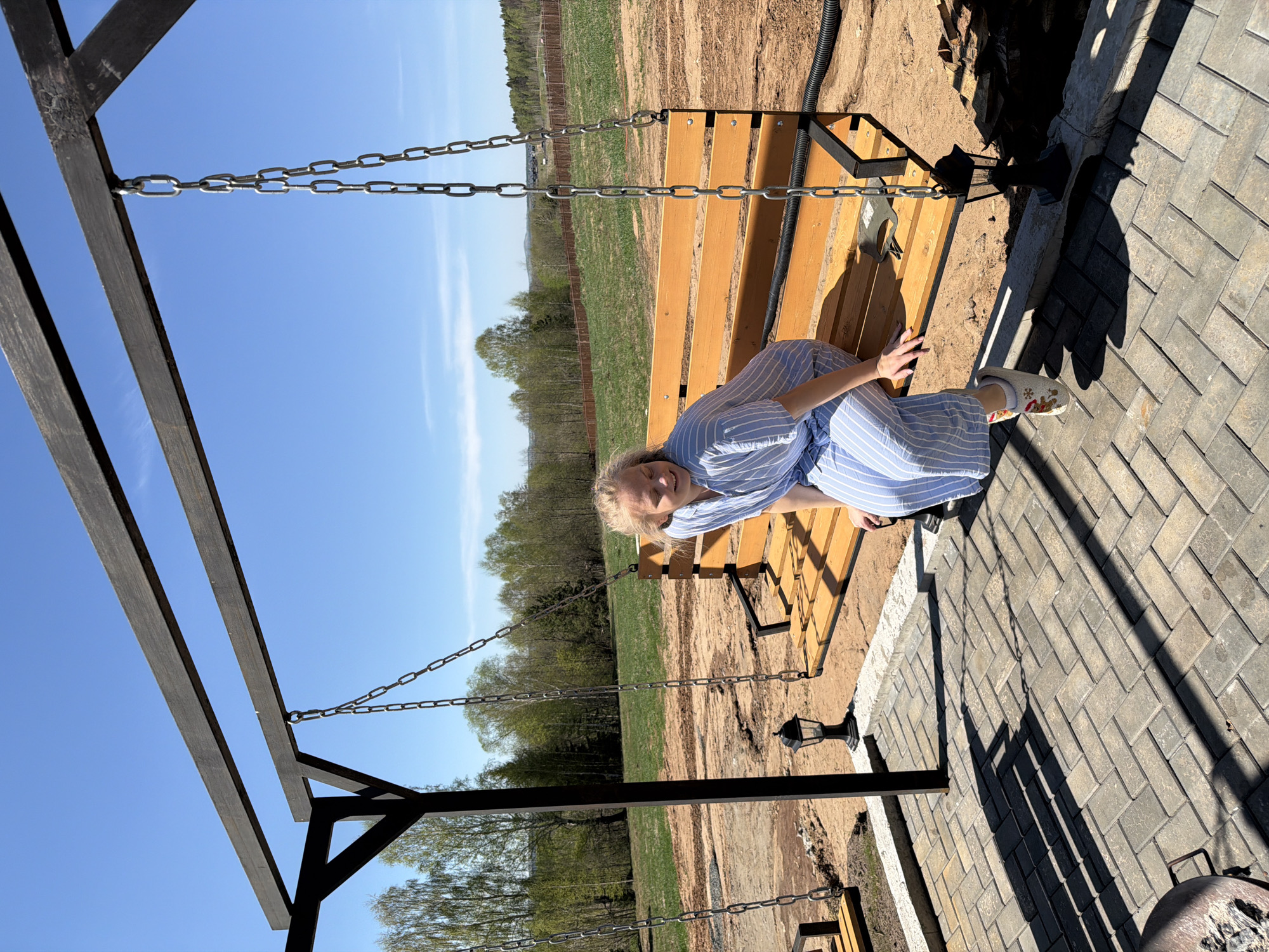 A woman sits on a swing bench in Kamennoye, Russia, on a sunny day. She wears a blue and white striped outfit and light-colored shoes. The swing bench is suspended by chains from a dark-colored wooden frame. The background includes a field and trees under a clear blue sky. A paved area with rectangular stones is visible adjacent to the swing.