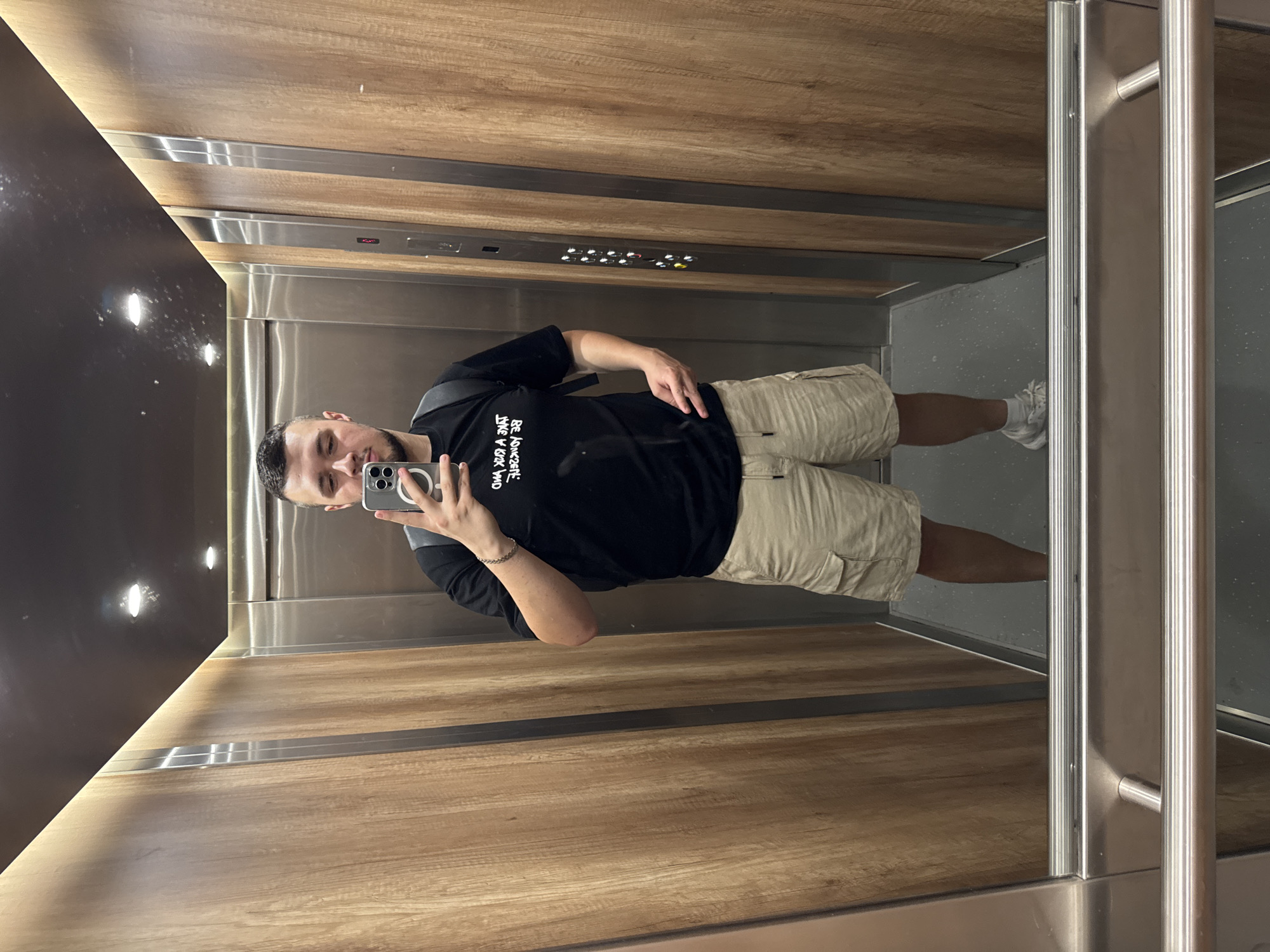 A man takes a selfie in an elevator. The elevator has wood-paneled walls, a dark ceiling with recessed lights, and a stainless steel control panel. The floor is gray. The man is wearing a black T-shirt, khaki shorts, and white sneakers. He is holding a smartphone in his left hand. The location is within the city of Sofia, Bulgaria.