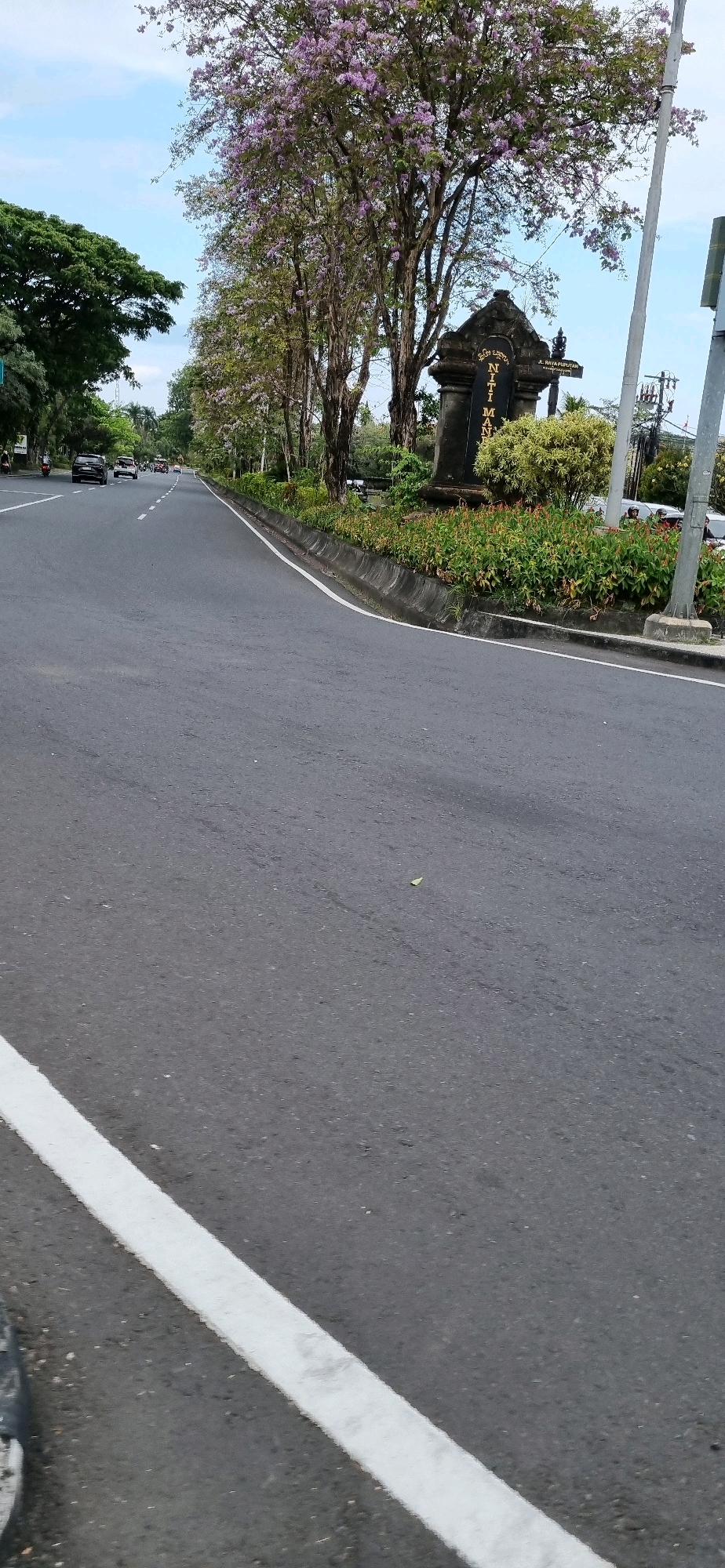 The image depicts a street scene in Denpasar, Indonesia, likely a main thoroughfare. In the foreground, a paved asphalt road is visible with a white line marking the edge of the lane. The road curves gently to the right. Along the right side of the road, there is a raised concrete curb bordering a median strip filled with lush green plants and some flowering red plants. Behind the median, a stone monument with inscribed text is present, partially obscured by a flowering tree with purple blossoms. The monument has a traditional Balinese architectural style.
Further down the road, several cars and motorcycles are in motion, indicating traffic. On the left side of the road, there are trees with dense green foliage. The sky above is a pale blue with some scattered clouds, suggesting it is daytime. The overall impression is that of a well-maintained urban road in a tropical environment. The inscription on the monument appears to include "NITI MANDALA," a common term in Bali referring to designated areas or administrative centers. The presence of the monument and the style of the surrounding vegetation strongly suggest this is a significant location within Denpasar.
