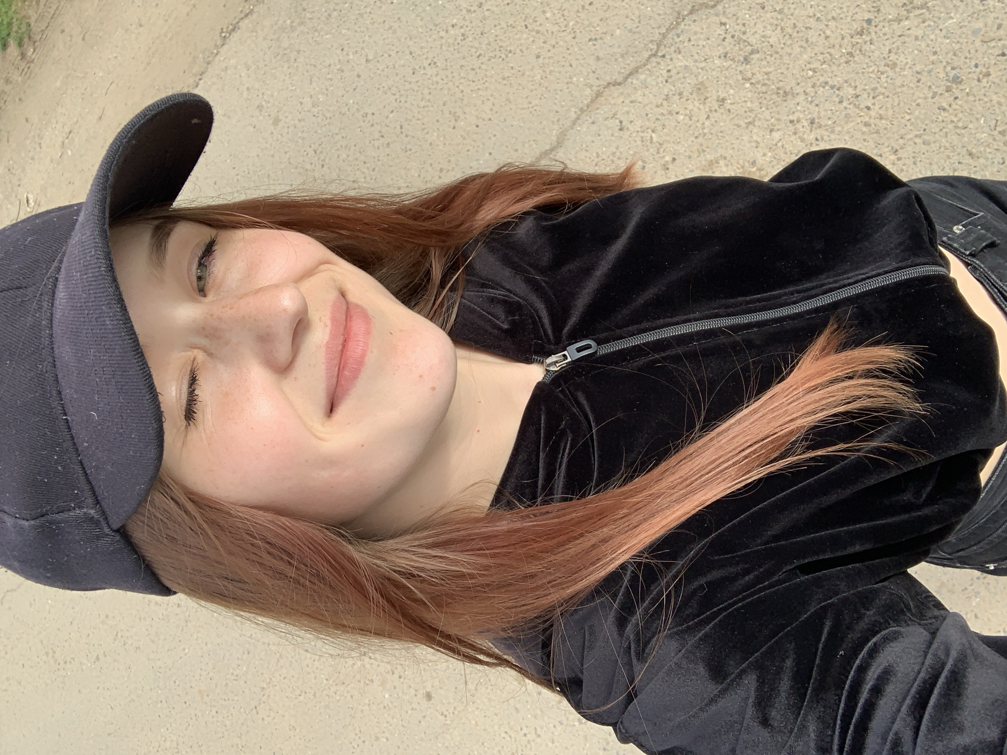 A portrait of a woman, presumably in her late teens to early twenties, with red-tinted brown hair, a black cap, and a black velvet jacket, is captured in an outdoor setting. She has freckles and her expression is one of gentle amusement, perhaps a slight smile. She is likely on a paved surface, possibly a sidewalk or road. The location information provided is Фоки, Russia; however, without more visual context within the image, this cannot be verified.