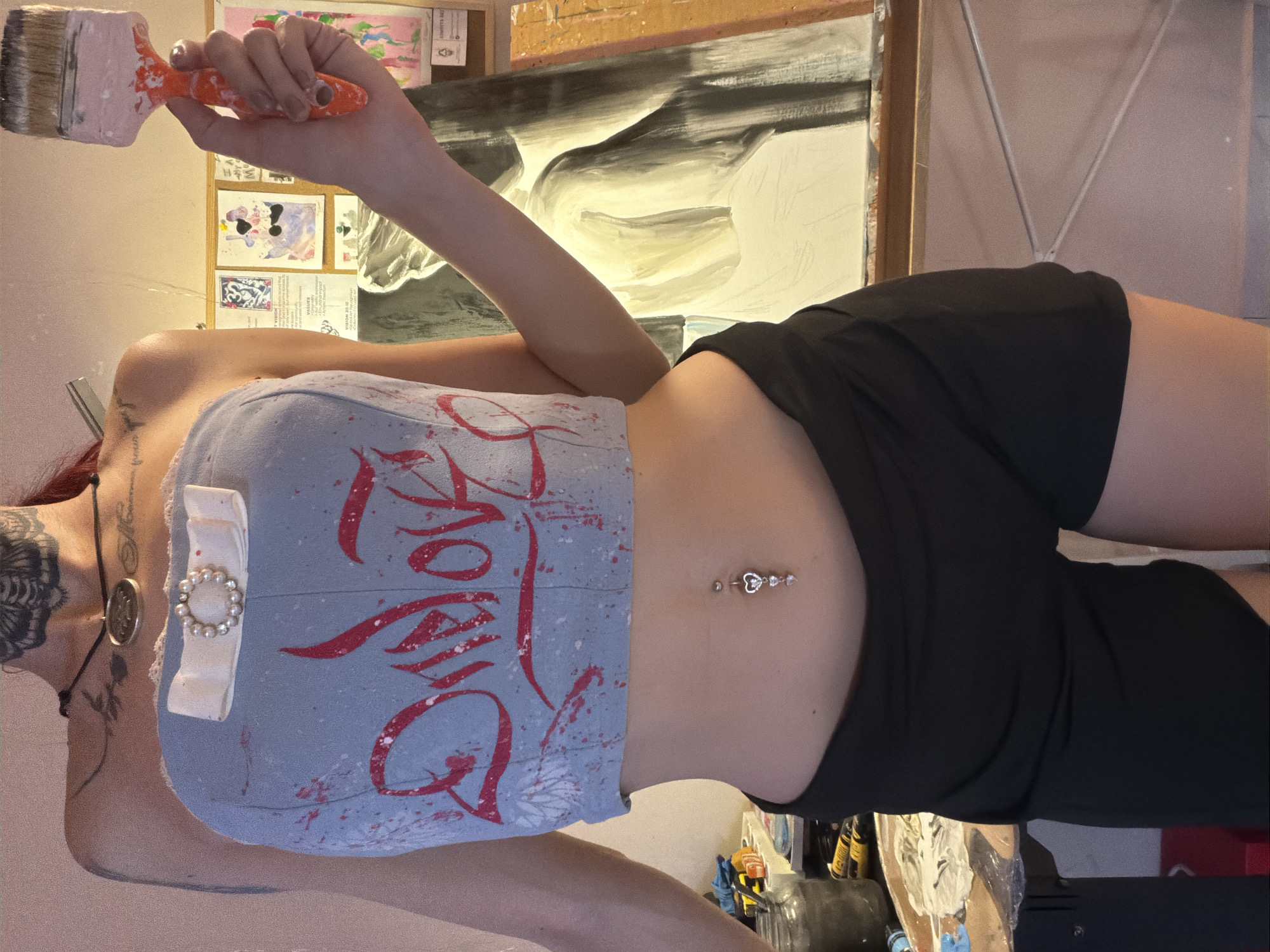 A person is standing in front of an easel with a partially finished painting. They are wearing a light blue strapless top with red graffiti and a white bow with pearls on the chest, and black shorts. The person is holding an orange-handled paintbrush with pink paint on it. The background shows a bulletin board with artwork and other papers. The scene appears to be an art studio. There is no information available that places this image in Svilengrad, Bulgaria.