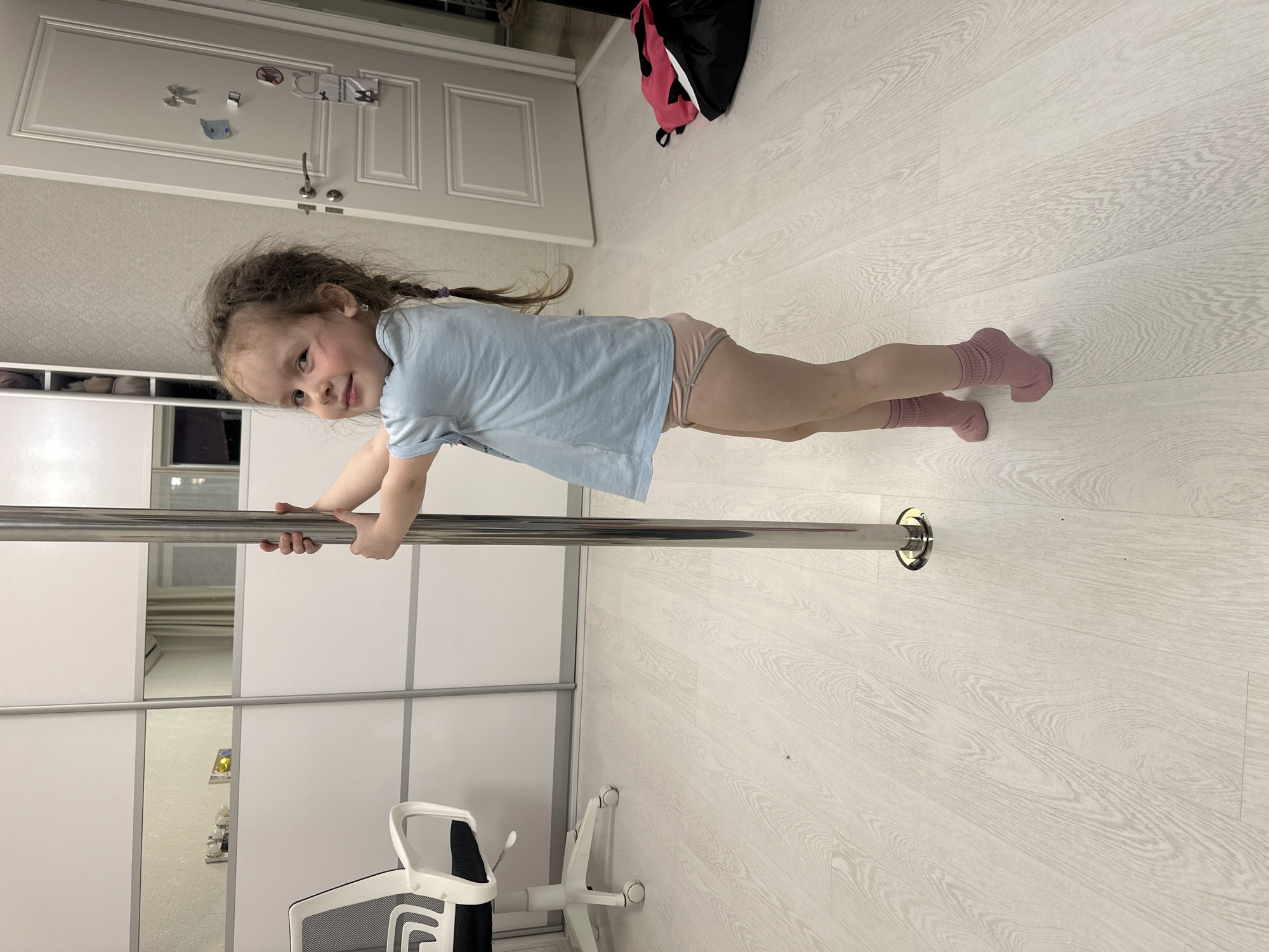A young girl is performing a plank exercise while holding onto a horizontal metal bar. She is wearing a light blue t-shirt and pink socks. The exercise is performed on a light-colored wooden floor in a room with white walls and a white closet with sliding doors. A white office chair is located nearby. A door can be seen in the background, and a black and pink bag is on the floor near the door. It is not possible to independently verify the city of Tomilino, Russia based on the provided image alone.