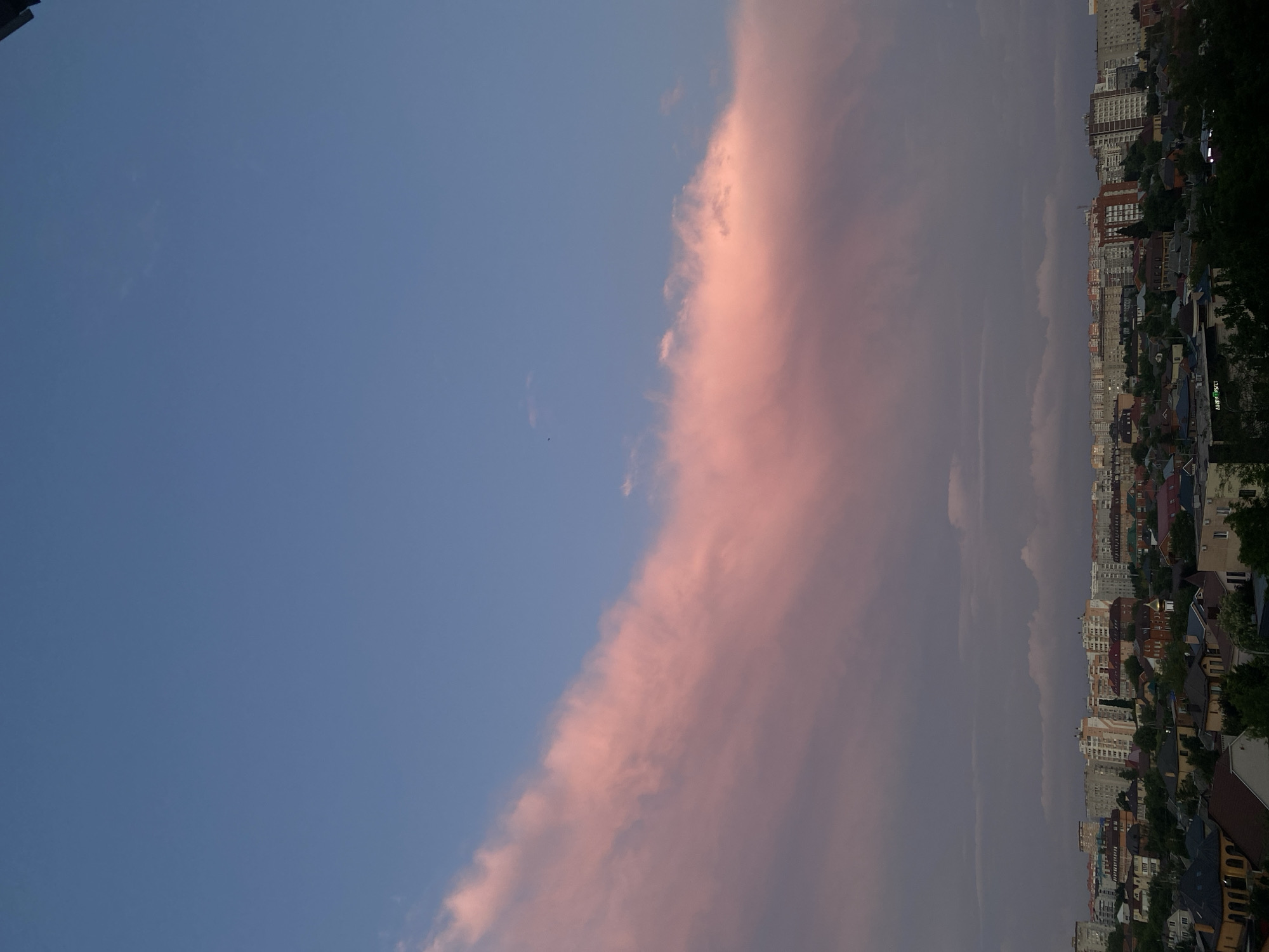 An aerial shot captures a city skyline under a dusky sky. The sky transitions from a clear blue at the top to a dramatic display of pink and grey clouds. A large, elongated pink cloud stretches diagonally across the mid-section of the frame, catching the last rays of the sun. Below the clouds, a dense urban landscape unfolds, characterized by a mix of modern high-rise buildings and smaller residential structures. Numerous rooftops are visible, some adorned with darker tiling, while others are lighter in color. Lush green trees are interspersed throughout the lower part of the scene, softening the hard lines of the city architecture. The overall impression is one of a city at dusk, with the sky providing a vibrant, atmospheric backdrop to the urban sprawl.