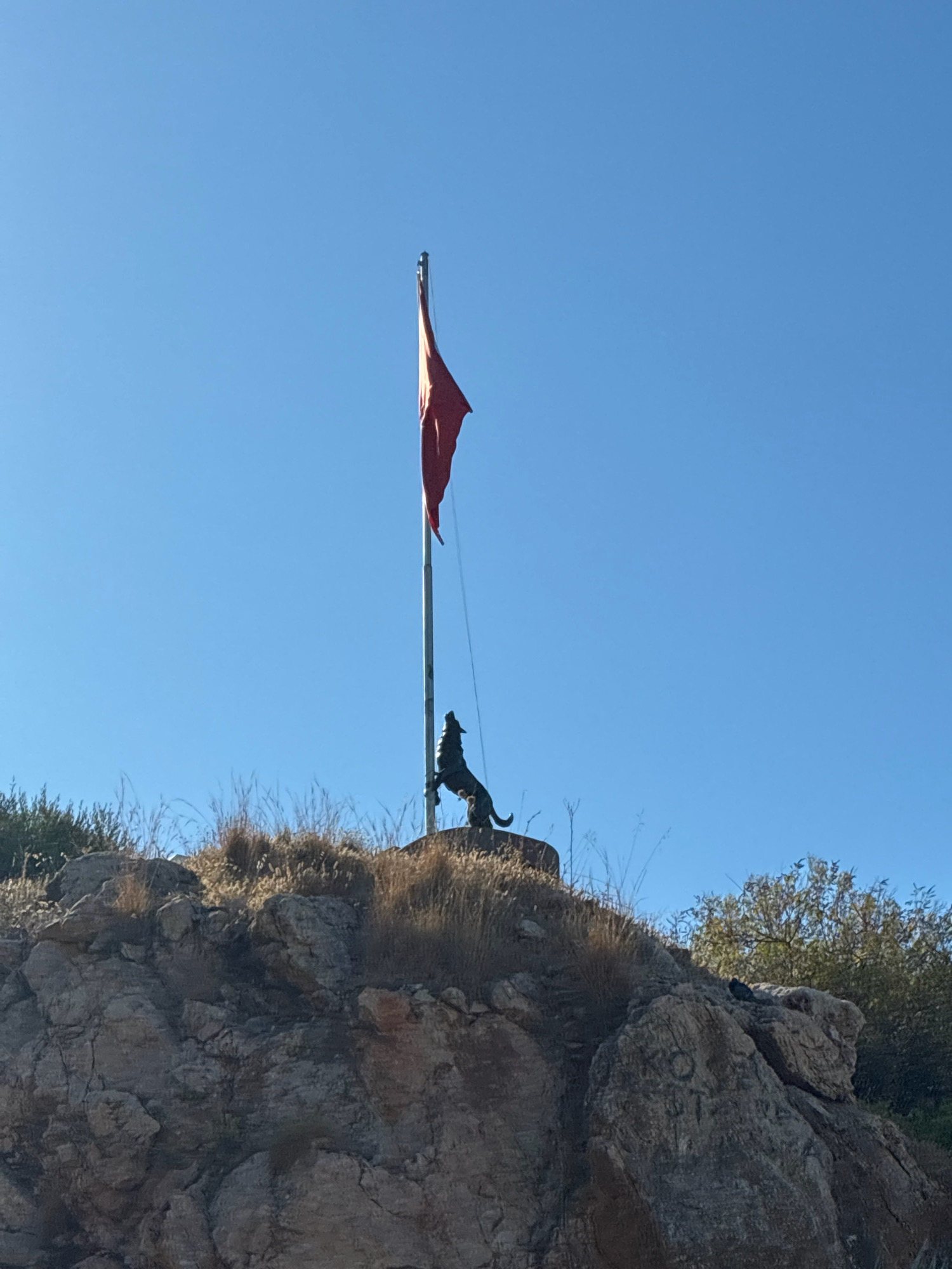 The image depicts a red flag on a flagpole, standing tall against a clear blue sky. At the base of the flagpole, a dark statue of a wolf stands with its head raised, as if howling. The flagpole and statue are positioned on a rocky outcrop covered with dry grass and shrubs. In the background, more rocky terrain and trees are visible. This monument, featuring a wolf and a flag, is located at the Atatürk Memorial in Alanya, Turkey, a historical site dedicated to Mustafa Kemal Atatürk, the founder of the Republic of Turkey.