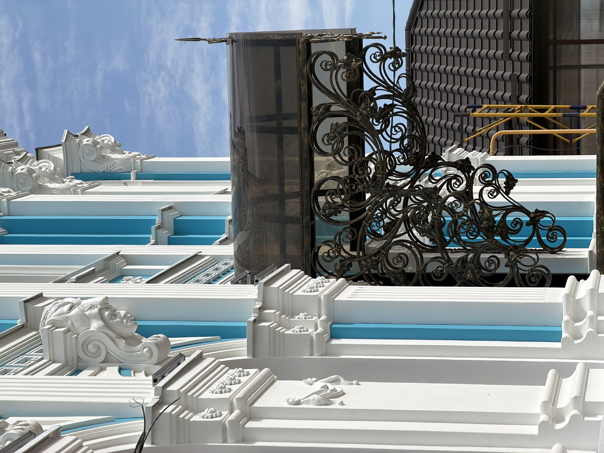 The image shows a portion of an ornate building with white columns and blue accents, located in Leningradskaya, Russia. The building features detailed white stucco sculptures, including faces on corbels, and decorative relief work. A dark, wrought-iron canopy with intricate scrollwork extends from the building, supported by brackets. Above the canopy, a segment of a brown tiled roof is visible. The sky above is blue with scattered clouds. A section of scaffolding with yellow and blue poles is positioned to the right of the building, partially obscured by the wrought-iron work and the roof. No people are present in the image, and no actions are depicted.