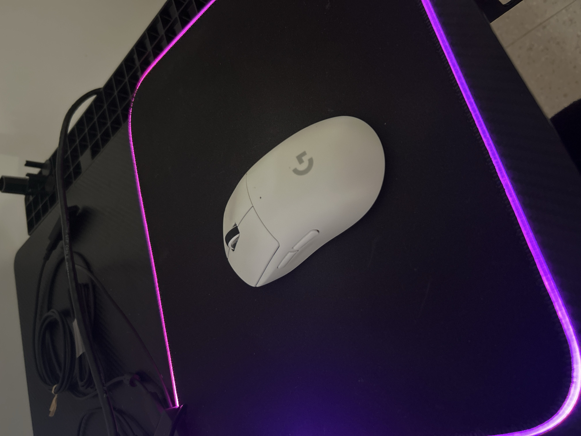 A white Logitech G Pro X Superlight mouse sits on a black gaming mouse pad with purple LED trim. The mouse is oriented towards the top of the frame, slightly off-center. The mouse pad is situated on a black desk or surface with a lattice-like structure visible along the left edge. Cables are bundled and connected to the device on the left. The location cannot be determined from this image.