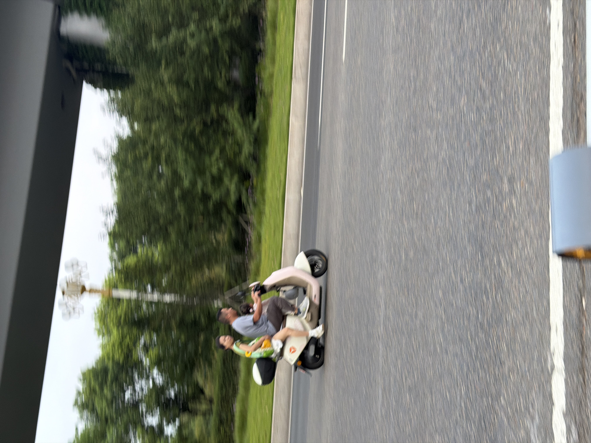 A father and his daughter are riding on a scooter. The father is driving, wearing a grey t-shirt and dark pants. His daughter is sitting in front of him, wearing a green dress. They are on a road in Jiangjin, China. The road is asphalt and has a white line on the side. There are trees and greenery in the background, with a cloudy sky above. The image is slightly blurred, suggesting motion.