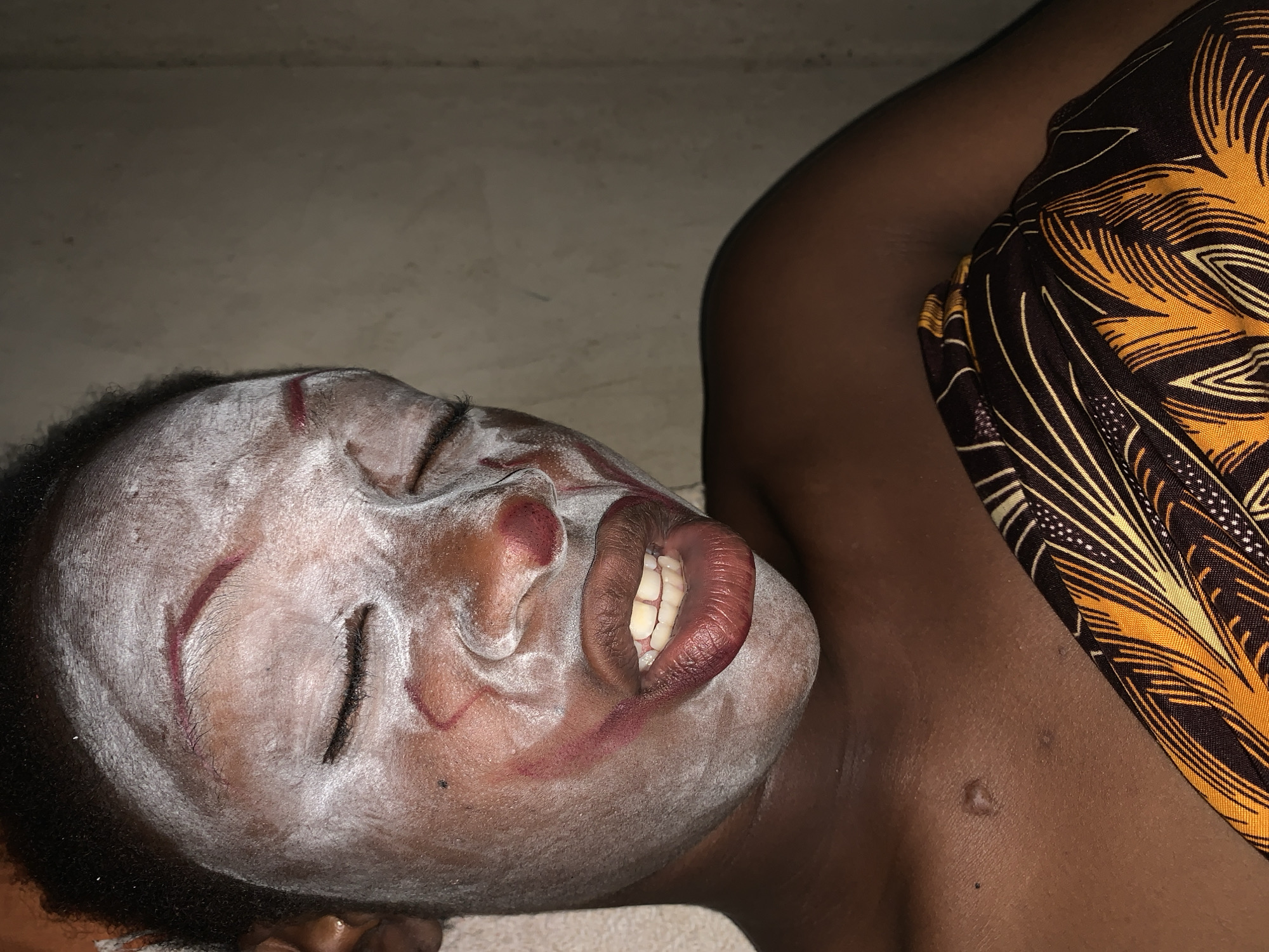 The image shows a person with dark skin lying on a light-colored surface. The person's face is covered in a white substance, with red lines drawn on their forehead and cheeks. Their eyes are closed, and their mouth is slightly open, revealing teeth. They are wearing a colorful patterned fabric draped over their shoulder. The location is identified as Agban, Nigeria.
