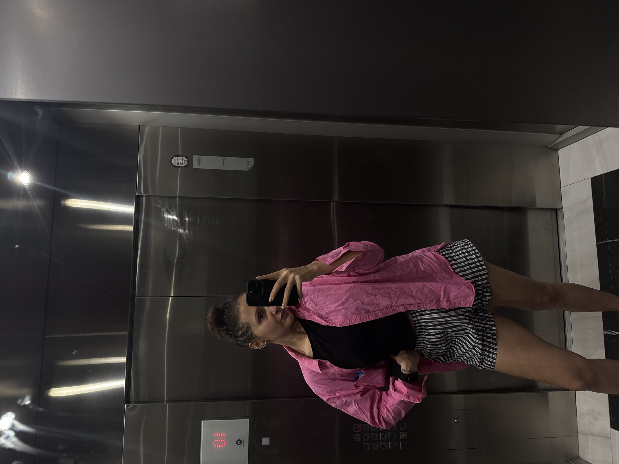 A woman is taking a selfie in an elevator. She is wearing a pink shirt, a black top, and striped shorts. The elevator has a stainless steel interior with buttons showing the floor numbers. The location is presumed to be Dubai, United Arab Emirates, based on image context clues.