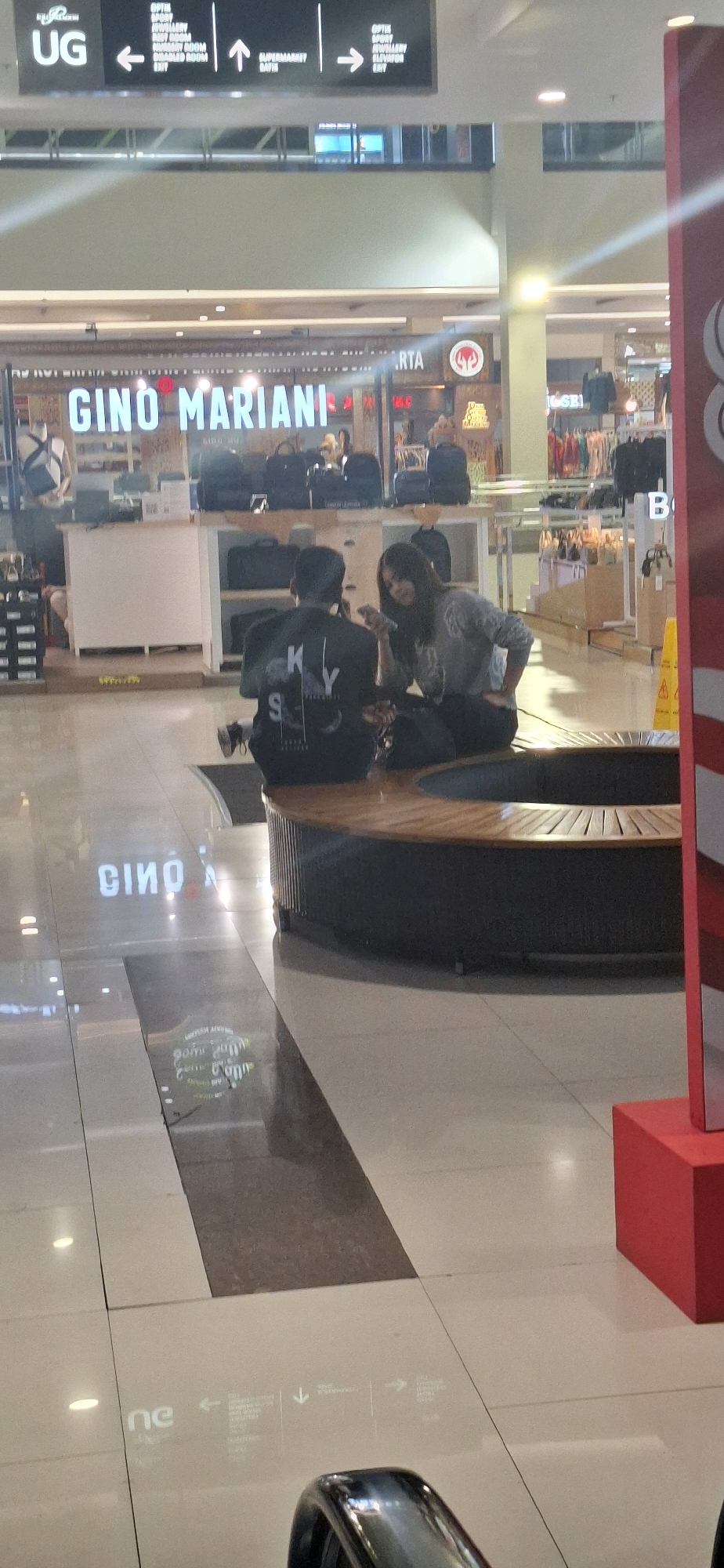 In Surakarta, Indonesia, inside a modern shopping mall, a young couple is seated on a circular wooden bench in the center of a spacious, brightly lit atrium. The man, facing away from the camera, wears a black t-shirt with white lettering. The woman, facing the man, is dressed in a gray long-sleeved top and dark pants, and she is looking at her phone. They are in the foreground, with a storefront featuring the "GINO MARIANI" logo prominently displayed behind them. The floor is polished and reflects the lighting and signage of the mall. The overall environment suggests a retail setting with shops and displays visible in the background.