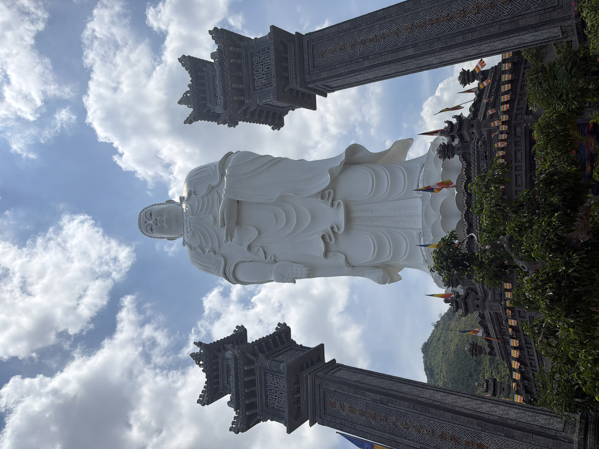 The image shows a low-angle view of a large, white Buddha statue in Tay Nha Trang Ward, Vietnam. The statue stands tall against a partly cloudy sky. On either side of the statue are decorative, dark gray pillars with intricate patterns. Behind the statue, foliage and other buildings are visible, suggesting a temple complex. Small colored flags are strung along the top of one building. A distant, green mountain is also visible.