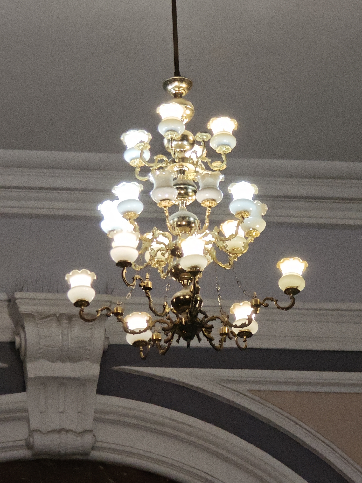 The image shows a close-up, head-on, eye-level view of an ornate, multi-tiered brass chandelier with numerous glass light fixtures. The chandelier is suspended from a dark rod against a neutral, textured ceiling with subtle architectural molding. The light fixtures, in the shape of inverted floral cups, emit a warm, yellowish light, illuminating the surrounding brass elements and casting a soft glow. The chandelier has a complex structure with curving arms and decorative detailing. Below the chandelier, architectural elements of a building are visible, including a white, fluted column base and curved, dark and light-colored wall sections, suggesting an interior space of a grand building. The image does not contain any people or indicate a specific location in Moscow, Russia.