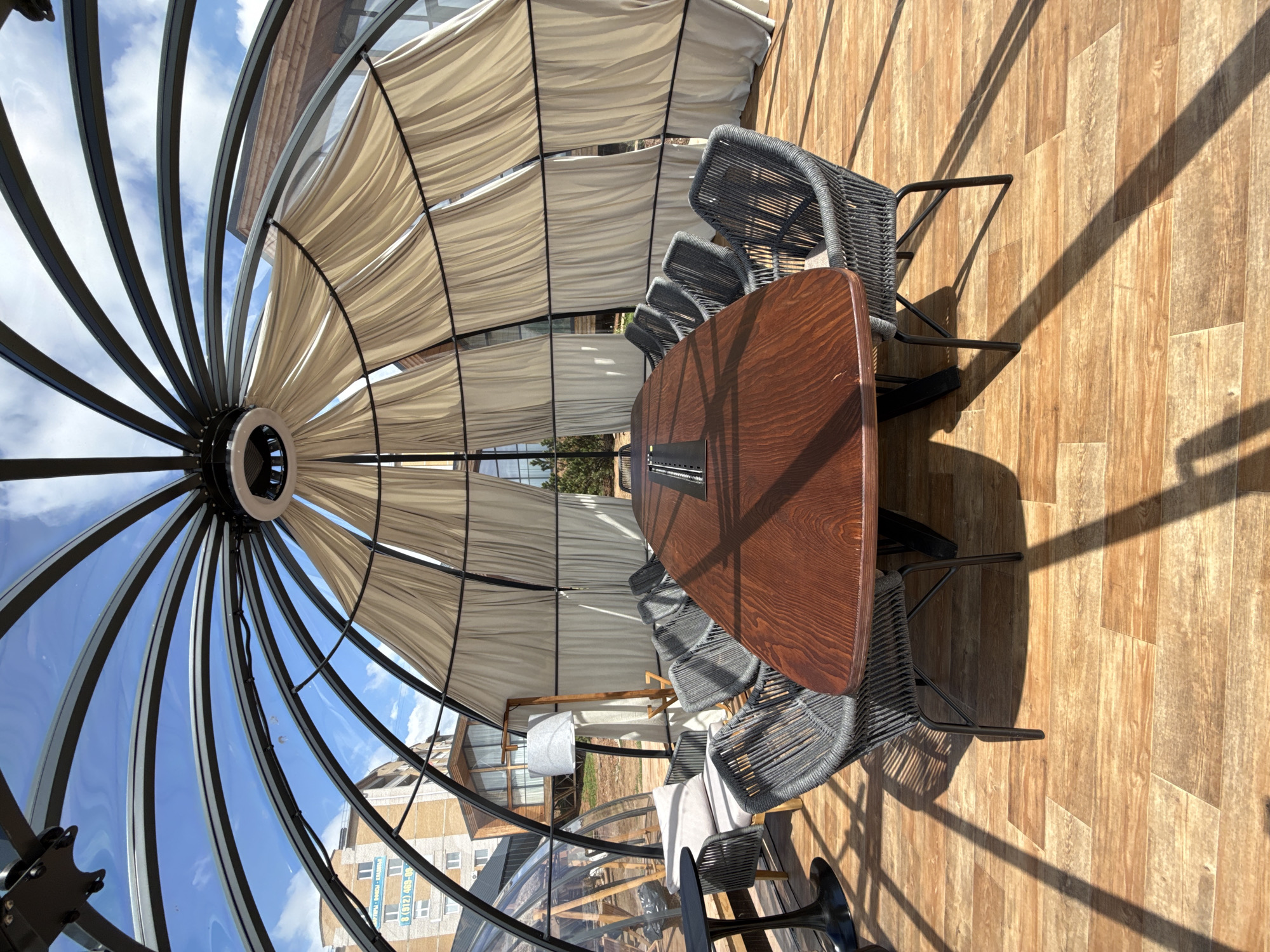 The image presents an interior view of a covered outdoor dining structure, possibly located in Votkinsk, Russia, based on the building visible through the structure's glass. The structure has a metal frame creating a dome-like enclosure with alternating panels of glass and white fabric. A large, oval-shaped wooden table dominates the scene, surrounded by gray woven chairs. Sunlight streams into the space, creating strong shadows on the wooden floor. The scene appears to be a restaurant or cafe setting.