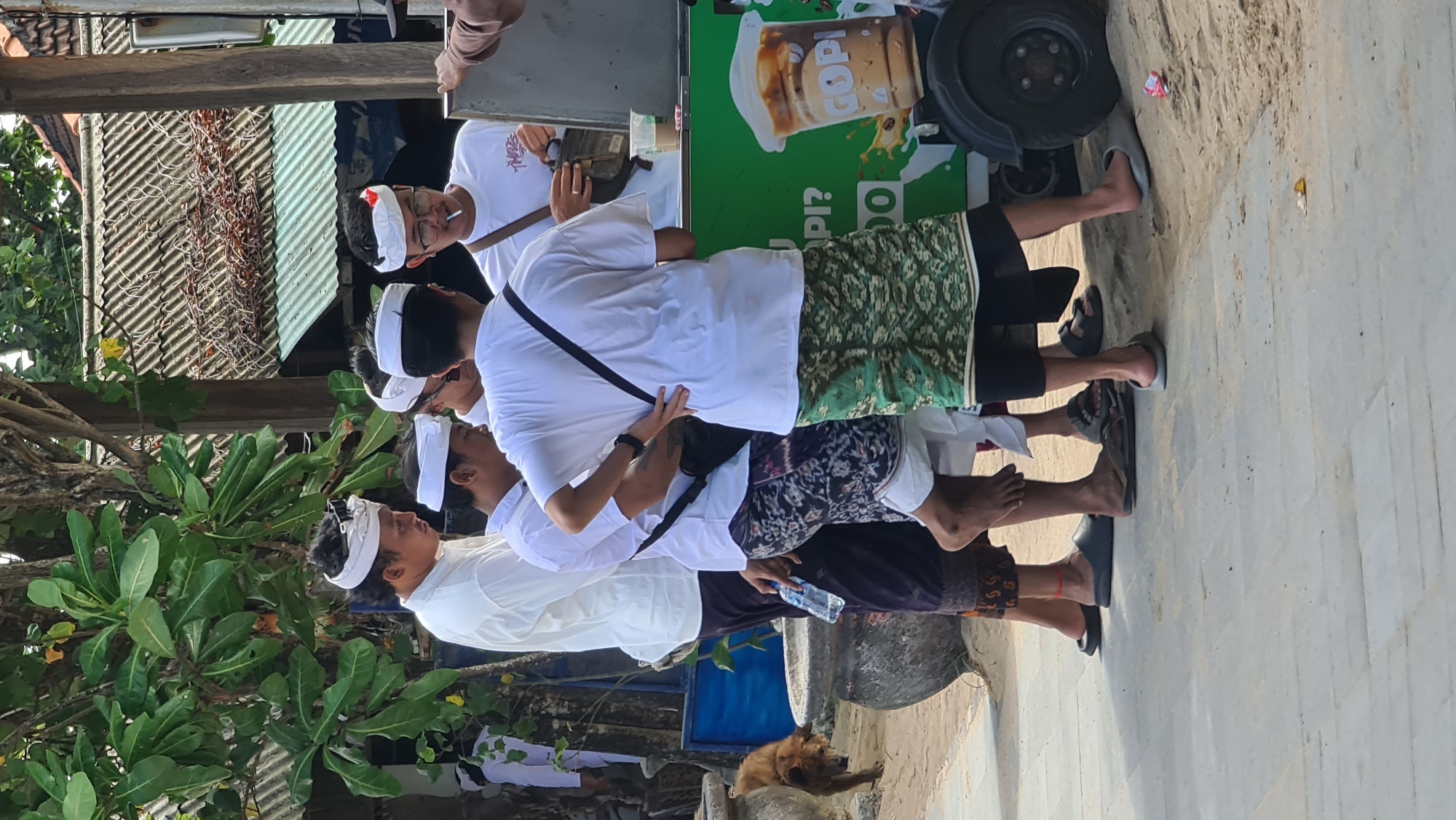 The image shows a group of men in traditional Balinese attire, specifically white shirts and sarongs, standing around a food cart. One man is holding a bottle of water. The food cart is green and has an advertisement for a coffee drink. The scene appears to be outdoors, with trees and a building in the background. There is also a dog in the lower left corner of the image. The presence of the food cart and the casual attire of the people suggest a public space, possibly a market or a tourist area. The architecture in the background, with the tiled roof, could indicate a location in Southeast Asia, and given the mention of Denpasar, Indonesia, it is likely a scene from that city. The overall impression is of a warm, sunny day in a bustling local environment.