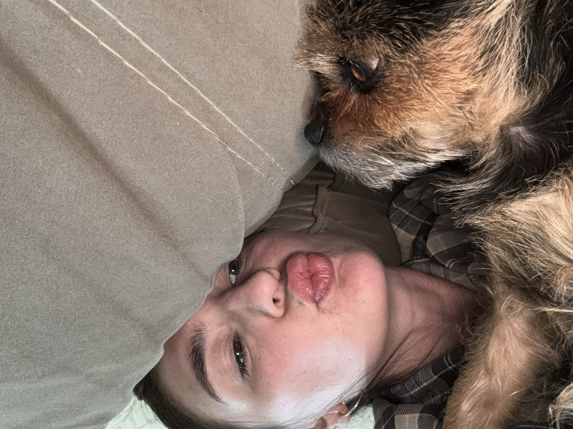 A woman is lying down with a dog next to her. She is partially covered by a beige blanket with two parallel seams. Her lips are puckered as if to kiss the camera. The dog, which is light brown and black, is nestled beside her. She is wearing a dark plaid shirt.
The location in the city of Sochi, Russia cannot be determined from the given image.