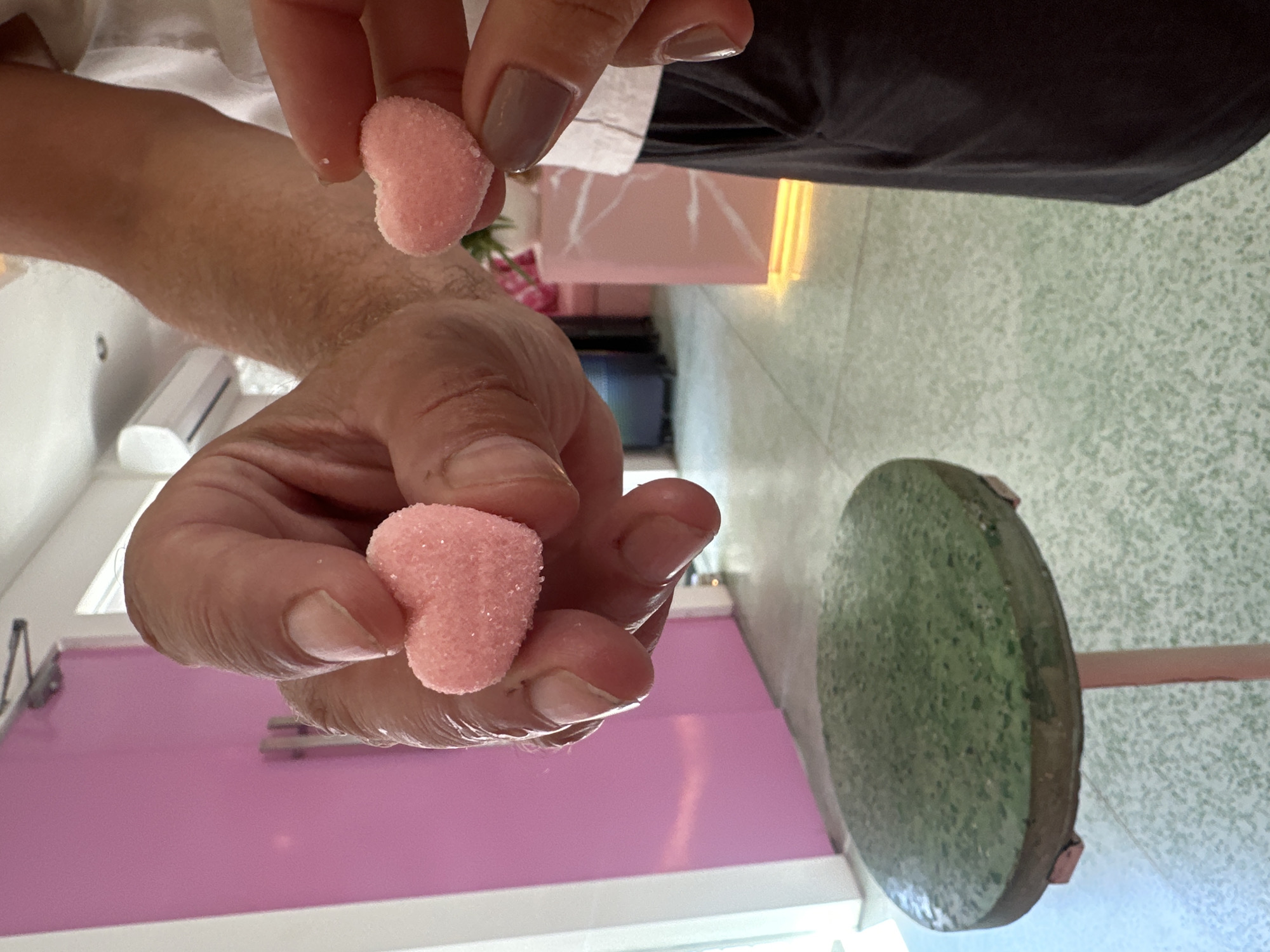 The image shows two people holding heart-shaped candies in their hands in what appears to be a dessert shop. The person on the left has fair skin and is holding a heart-shaped candy in their right hand. The person on the right has light-tan skin and is holding a heart-shaped candy in their left hand. The shop has pink walls and furniture. There is a green stool in the foreground. The location is North Kuta, Indonesia.