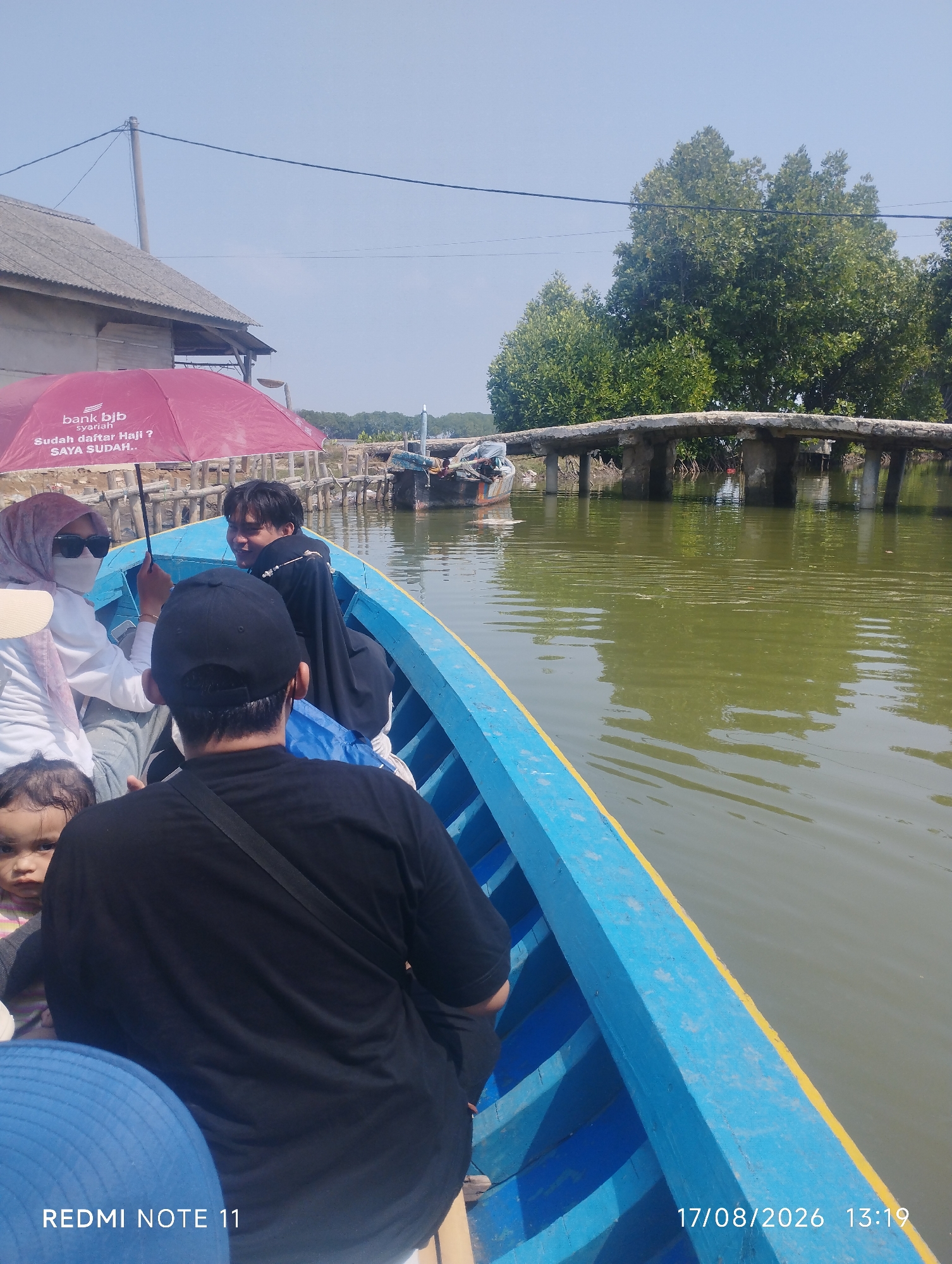 The image depicts a group of people on a blue wooden boat navigating a body of water, likely a river or canal, under a clear sky. The boat is positioned from a first-person perspective, looking forward. In the boat, a woman wearing a hijab and sunglasses is seated, holding a red umbrella with "bank bjb syariah" and Indonesian text visible. A young child with dark hair is also visible in the boat, looking towards the camera. A man wearing a black t-shirt and a black baseball cap is in the foreground, facing away from the camera. Another person in the boat, a young man, is looking back towards the rear of the boat.
The environment surrounding the water includes a rustic building with a gray roof to the left, adjacent to a wooden structure. Dense green foliage, likely mangrove trees, lines the opposite bank. A concrete bridge with visible supports spans the water in the mid-ground. Several other boats are moored or visible further down the waterway. The water itself appears murky green and reflects the surrounding trees and sky. A timestamp and the text "REDMI NOTE 11" are overlaid on the lower portion of the image, indicating it was taken on August 17, 2026, at 13:19. This scene is consistent with a location in Karawang, Indonesia, possibly a coastal or riverine area known for its mangrove forests and local transportation by boat.