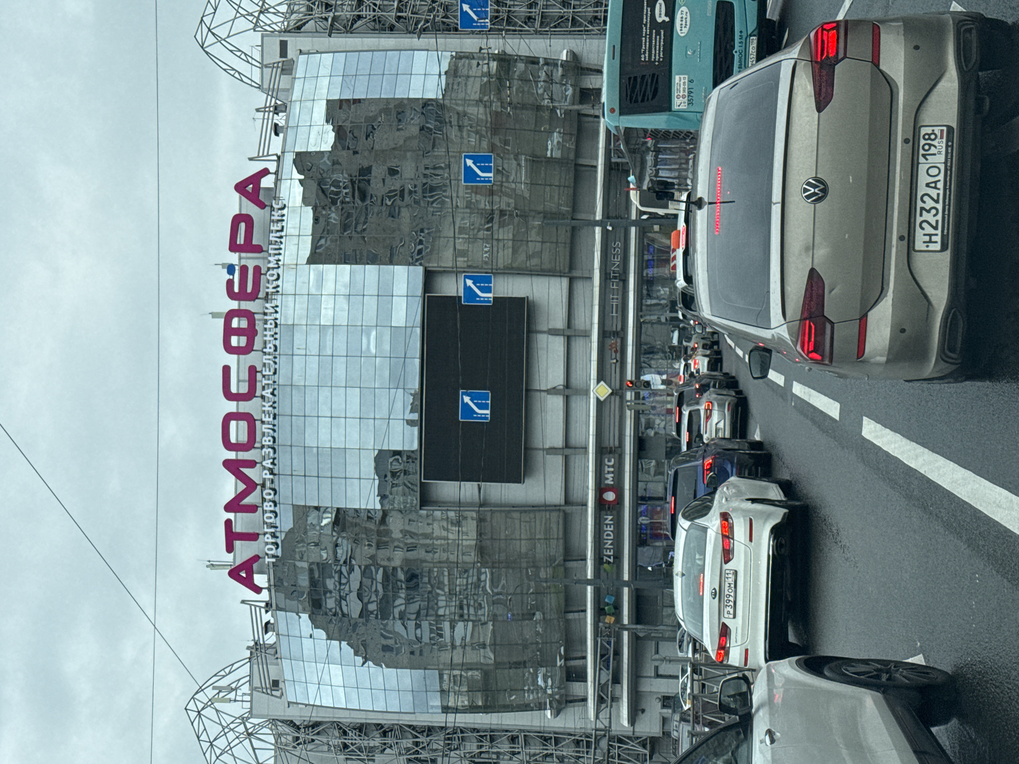 The image depicts a traffic jam in Saint Petersburg, Russia, adjacent to the "Atmosfera" shopping and entertainment complex. Several vehicles are visible, including a Volkswagen with license plate H232AO 198 and a Kia with license plate P399 OM 11. Traffic direction signs are also visible. Storefront signs for "Zenden," "MTC," and "Hit Fitness" can be identified. The building is made of glass and steel. The sky is overcast.