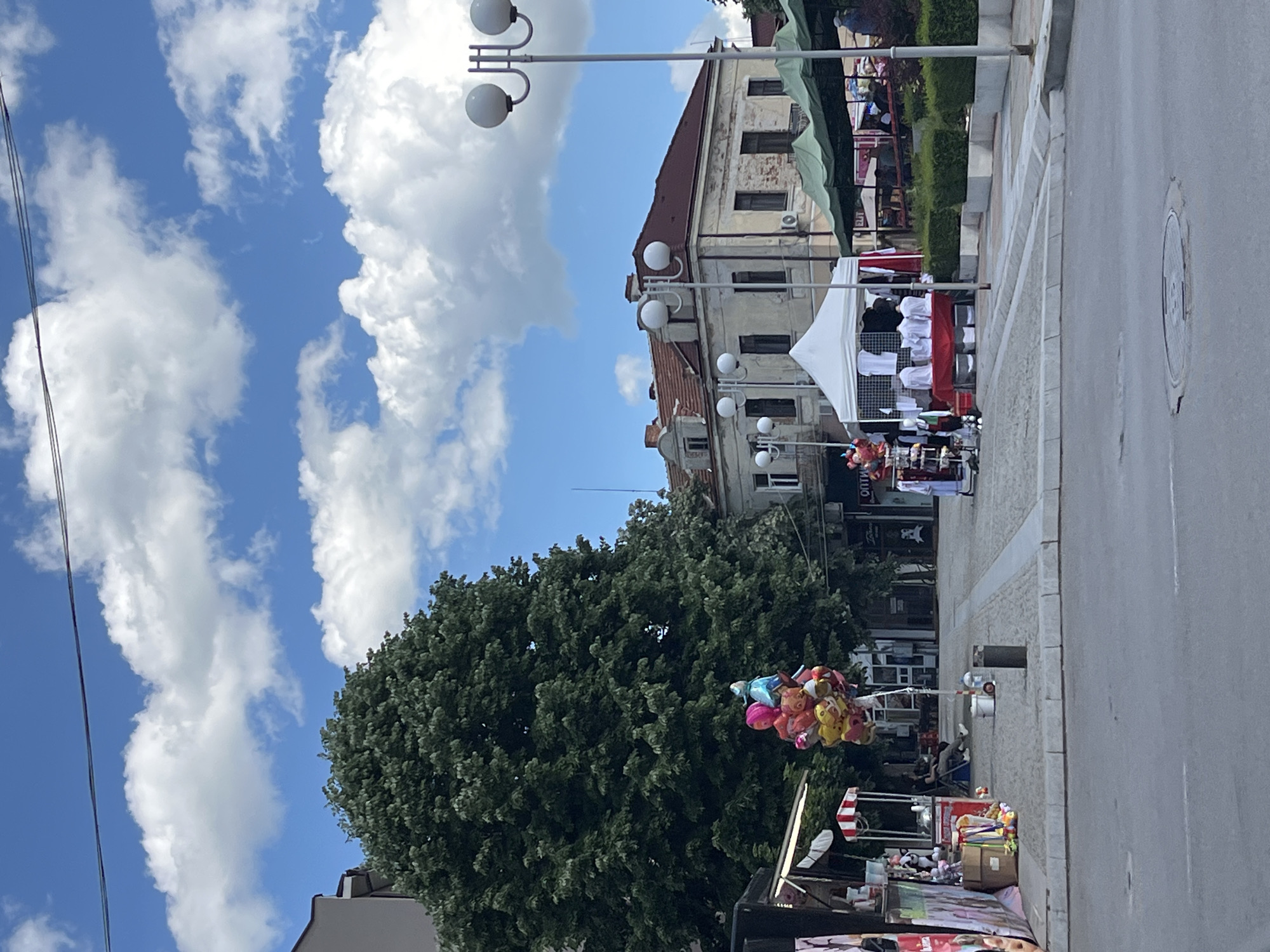 The image depicts a street scene in Elhovo, Bulgaria, marked by a vibrant sky with scattered cumulus clouds. A large, mature tree stands to the left, partially obscuring an older, multi-story building with a red-tiled roof. The building features multiple windows and appears to be constructed of aged stone or concrete.
The street is lined with vendors selling various items, including balloons held by a cart and a stand covered with white shirts under a white tent. Other merchandise includes what appears to be toys. Light poles with spherical lamps are a recurring feature along the street. The foreground shows an asphalt road with a manhole cover. People can be seen walking and sitting along the street.