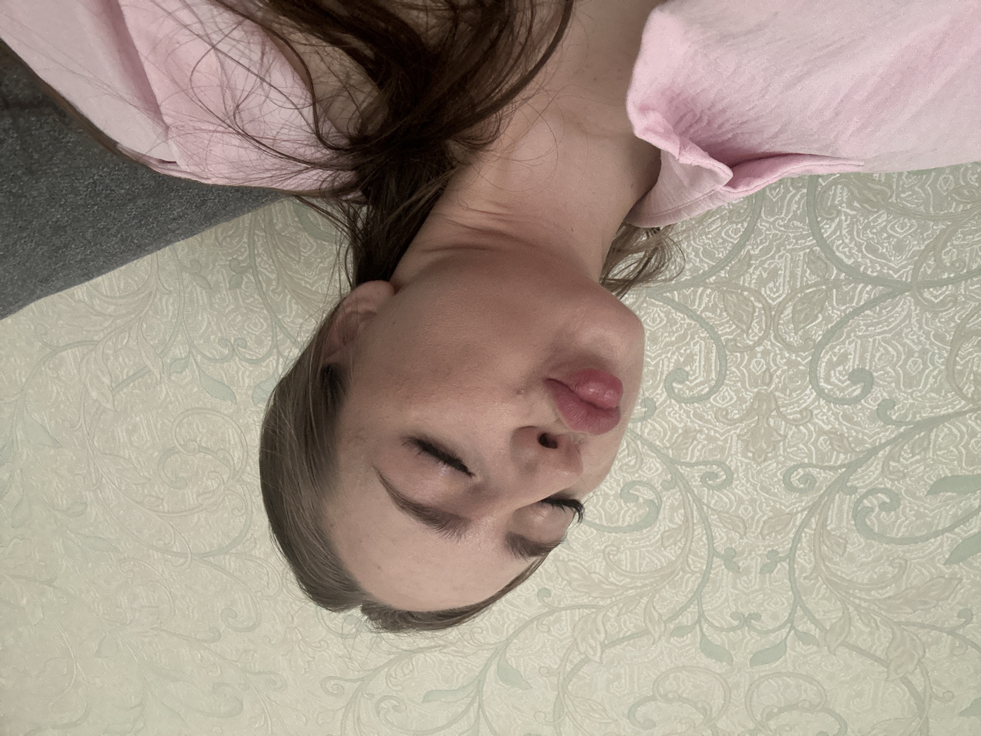 A young woman with long brown hair and closed eyes puckers her lips in a selfie. She is wearing a pink collared shirt and is positioned in front of a wall with a light green and beige patterned wallpaper. A dark gray upholstered surface is visible in the right portion of the image. The scene appears to be indoors.
