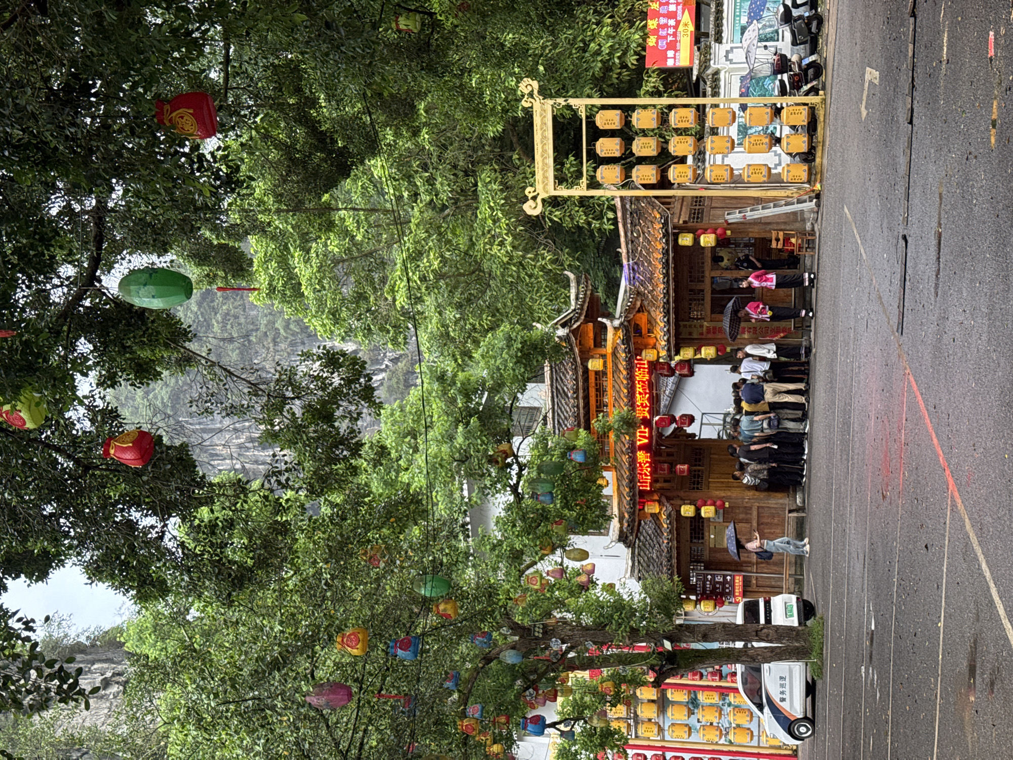 The image depicts a scene in Guilin, China. In the foreground, trees with brightly colored lanterns hanging from their branches frame the view. Beyond the trees, a traditional Chinese building with a tiled roof and red lanterns is visible. A gold, rectangular structure with a grid of yellow boxes stands next to the building. A group of people is standing in front of the building. A woman holds an umbrella while walking in a parking lot where a small white vehicle is parked. The road is wet, suggesting recent or ongoing rain. In the background, a misty mountain landscape can be seen.