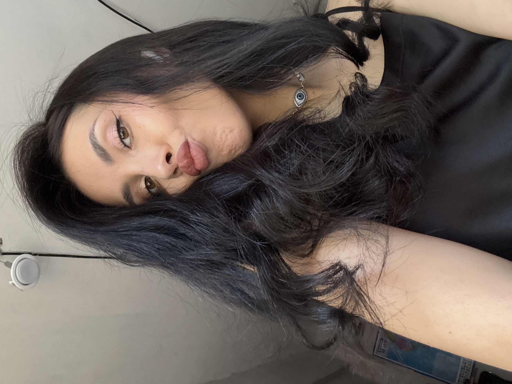 A close-up, rotated selfie features a young woman with long, dark, wavy hair that frames her face and flows over her shoulders. She has dark eyes, accentuated by light pink eyeshadow and winged black eyeliner. Her full lips are formed into a distinct pout, and she looks directly at the camera with a playful expression. A subtle crease or dimple is visible on her left cheek.
She wears a necklace with a small, eye-shaped pendant featuring a blue iris, resembling an "evil eye" charm. Her attire includes a simple black top, with a thin strap visible on her right shoulder. The background consists of a light-colored wall, indicating an indoor setting. A white circular fixture, possibly a light, and a black cord are visible in the upper left of the frame. A blurry object with blue and white elements, potentially a book or container, is partially seen in the lower right. The lighting is soft and even. No specific time of day or weather can be determined, and no text is visible within the image. The location context provided is Votkinsk, Russia.