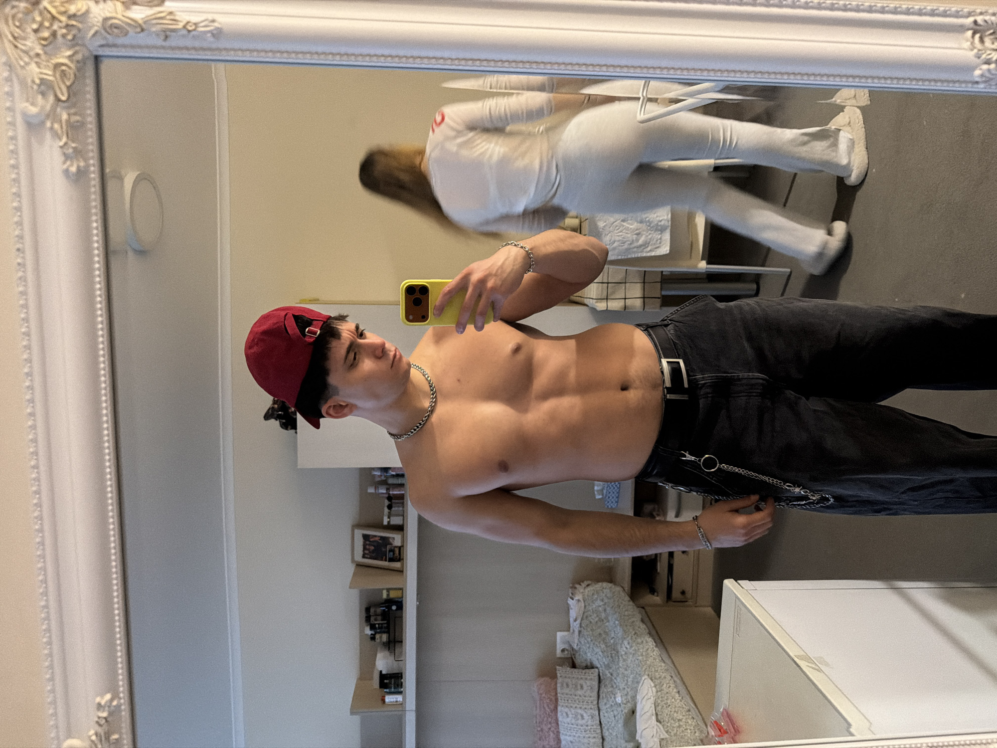 A shirtless man with a red cap takes a mirror selfie in a room with a white ornate mirror. He is wearing dark jeans with a chain. In the reflection, a woman wearing white athletic clothes is moving across the background. A white cabinet is visible in the lower right corner. A shelf with various items is seen behind the man. The location cannot be determined from the visual information provided.