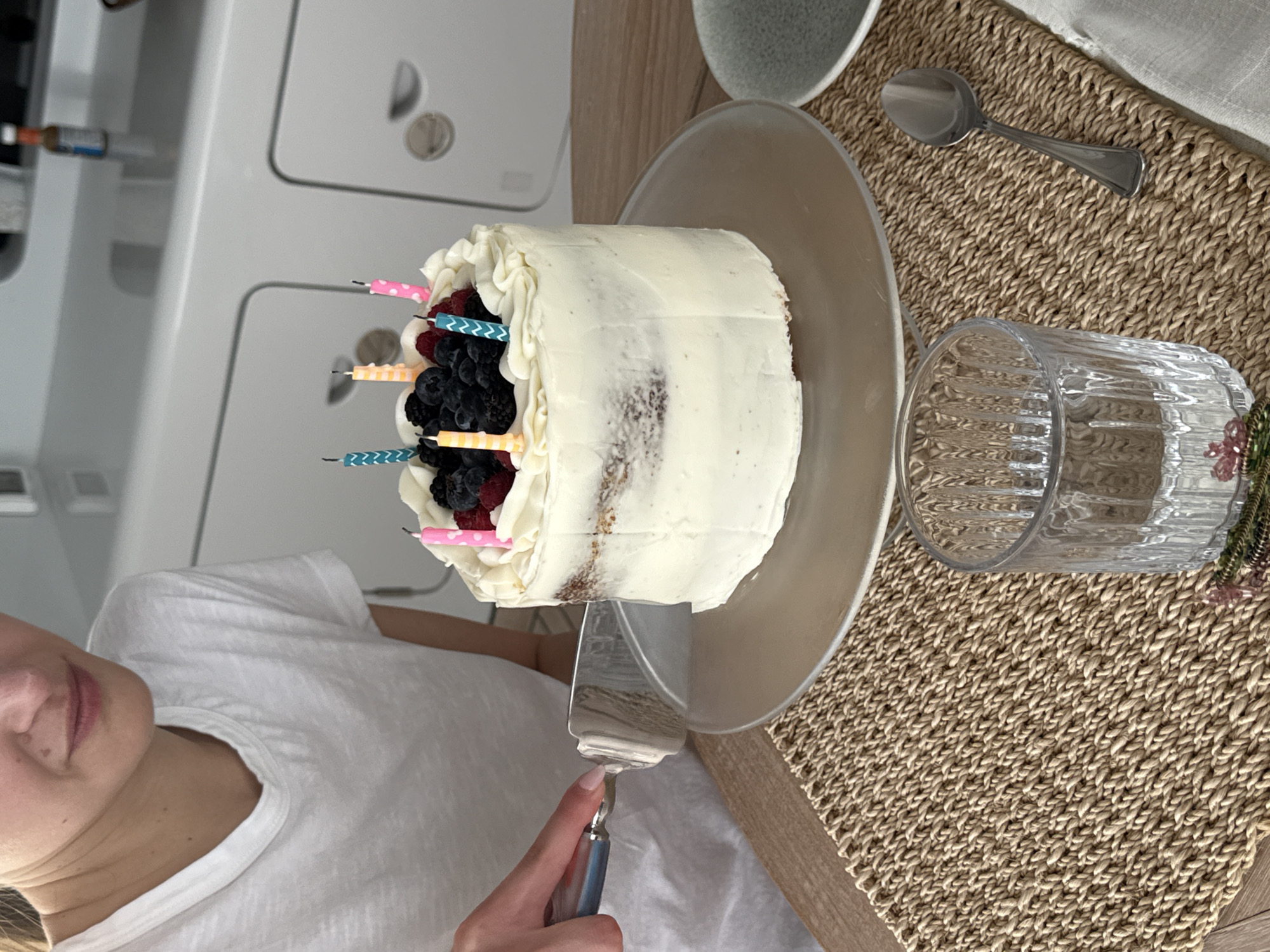 The image shows a person cutting a cake with a cake server. The cake is frosted with white icing and topped with fresh berries and birthday candles. It sits on a clear glass cake stand. A glass of water, a spoon, and a ceramic bowl are on a woven placemat next to the cake. The location is not specified.