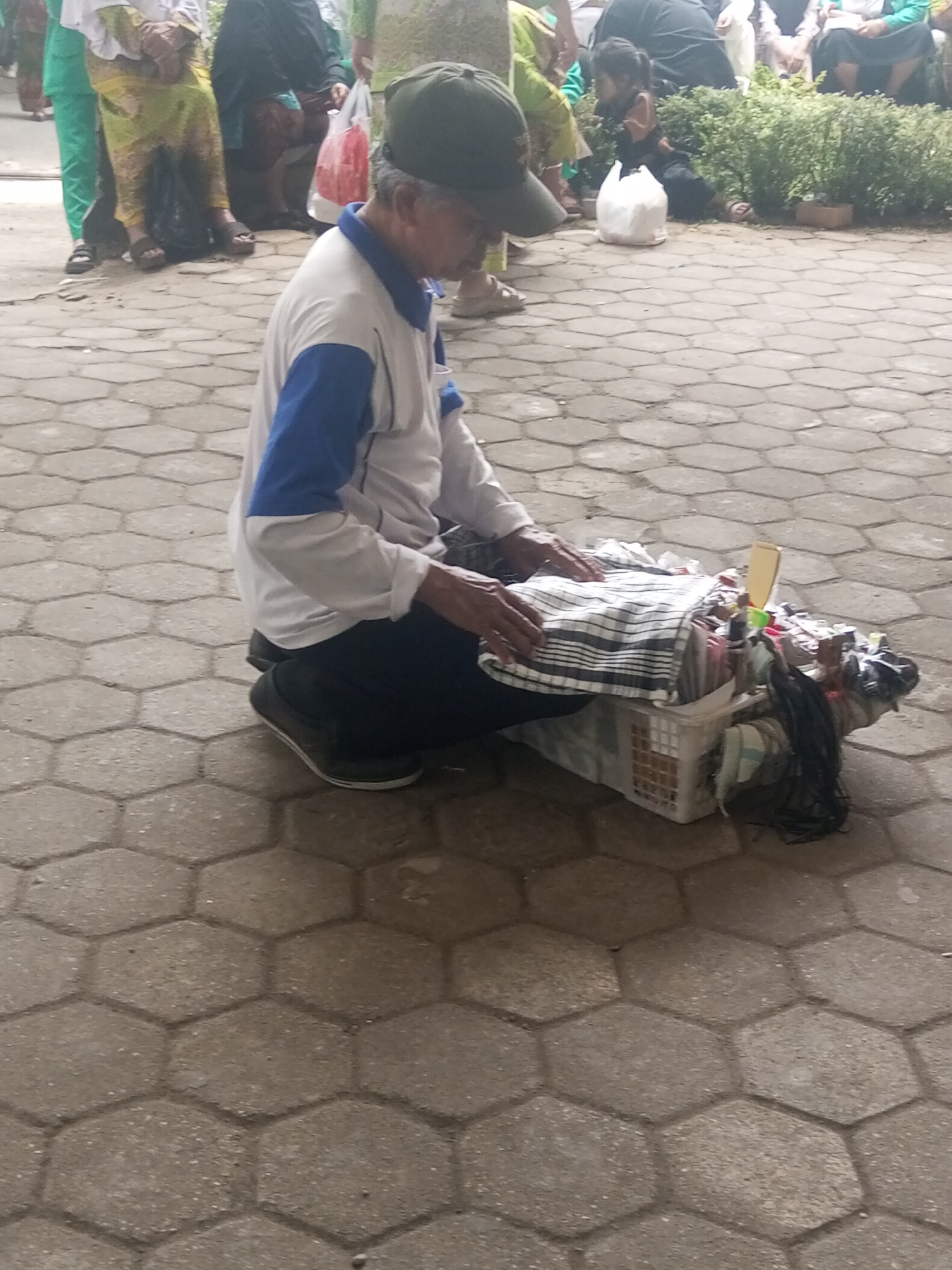 In Klaseman, Indonesia, a man wearing a green cap and a blue and white shirt kneels on a hexagonal paved surface. He is positioned in front of a white woven basket filled with assorted items, including fabrics, toiletries, and other small objects. His hands are resting on the fabrics in the basket. In the background, several people are visible, some of whom appear to be sitting or standing, and one child is seated near a white plastic bag amidst green foliage. The environment suggests an outdoor public space, possibly a market or a street.