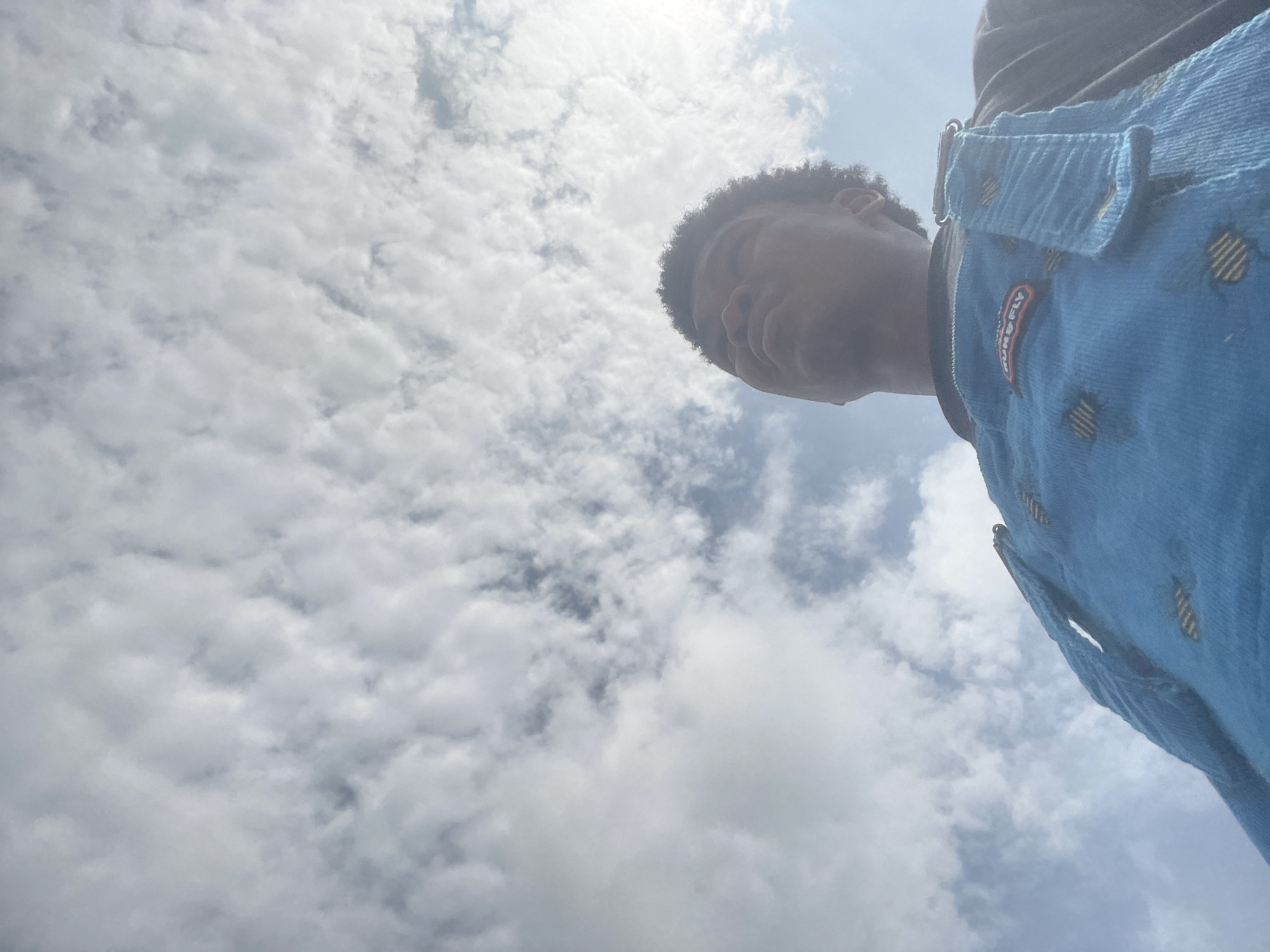 A low-angle, close-up, outdoor photograph shows a person wearing blue overalls under a bright, cloudy sky in Ilado, Nigeria. The person's face, shoulders, and the bib of the overalls are visible. The sky is filled with white, puffy clouds. The overalls are made of corduroy and have a patch with text that reads "RUN & FLY". The bottom of the image is predominantly the blue overalls, with the person's face and the sky occupying the upper two-thirds of the frame.