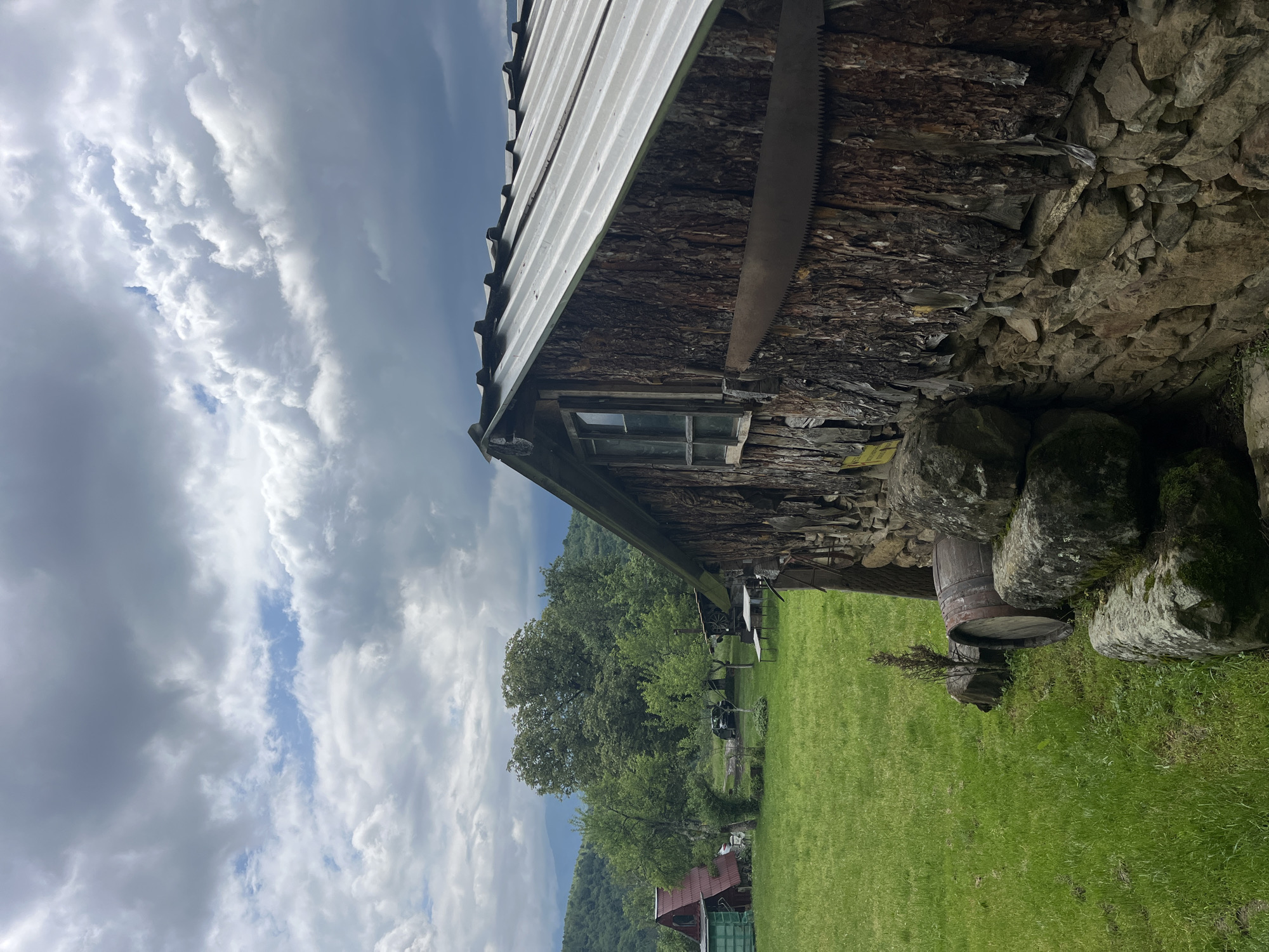 The image depicts an exterior view of a rustic structure located in Shopite, Bulgaria. The building appears to be constructed using a combination of materials including wood bark and stones. A metal sheet roof covers the structure. A weathered crosscut saw is mounted on the side of the building. A small window is visible near the top of the wooden section. The building is surrounded by a grassy area. A barrel sits near the base of the building, surrounded by rocks. In the background, there are trees and a structure with a red roof. The sky is filled with clouds, suggesting possibly inclement weather.