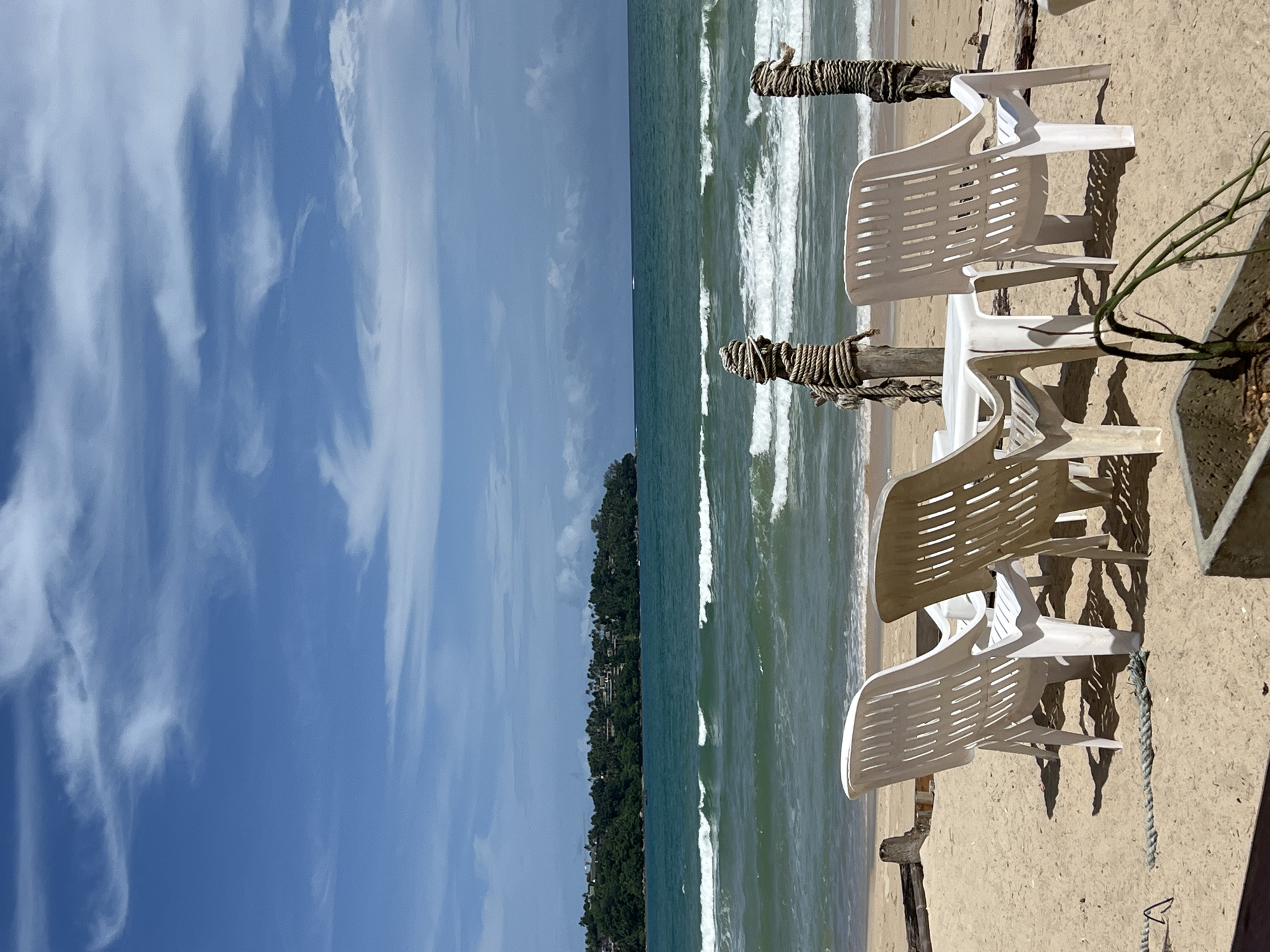The image depicts a serene beach scene with a view of the ocean under a partly cloudy blue sky. In the foreground, three white plastic chairs and a small white plastic table are arranged on the sand, suggesting a seating area for beachgoers. Two wooden posts, wrapped with thick rope, stand behind the table and chairs, adding a rustic touch to the setting. The beach is made of light-colored sand, with some debris and a piece of driftwood visible. The ocean is a vibrant turquoise green, with gentle waves rolling towards the shore. In the background, a lush green, tree-covered headland juts out into the sea, dotted with buildings. The overall impression is one of a peaceful and inviting tropical getaway.