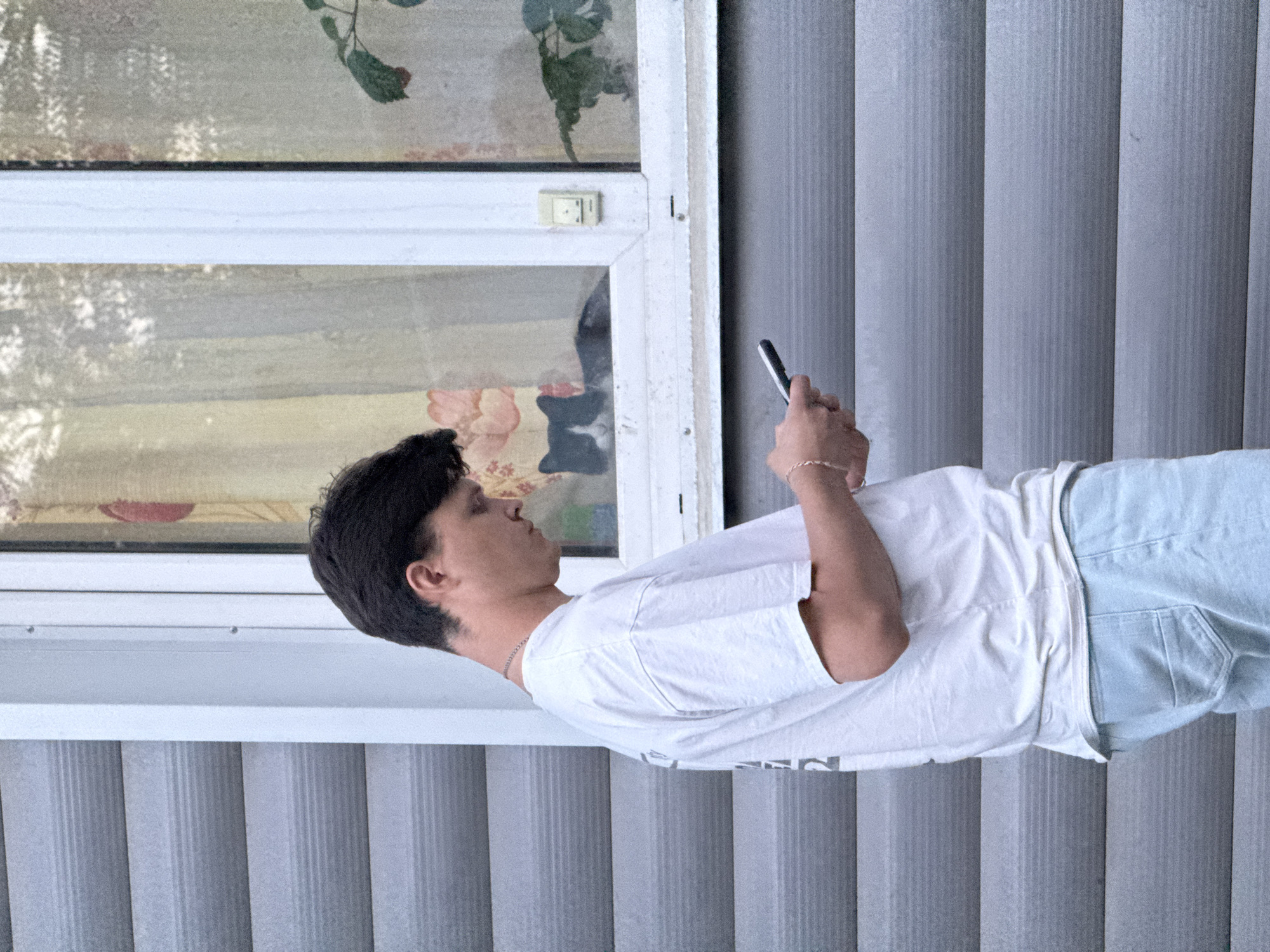 A person is standing indoors, looking at a mobile phone. The person is wearing a white t-shirt and light blue jeans. The background consists of a window and vertical blinds. The location cannot be determined as Ягул, Russia with the provided information.